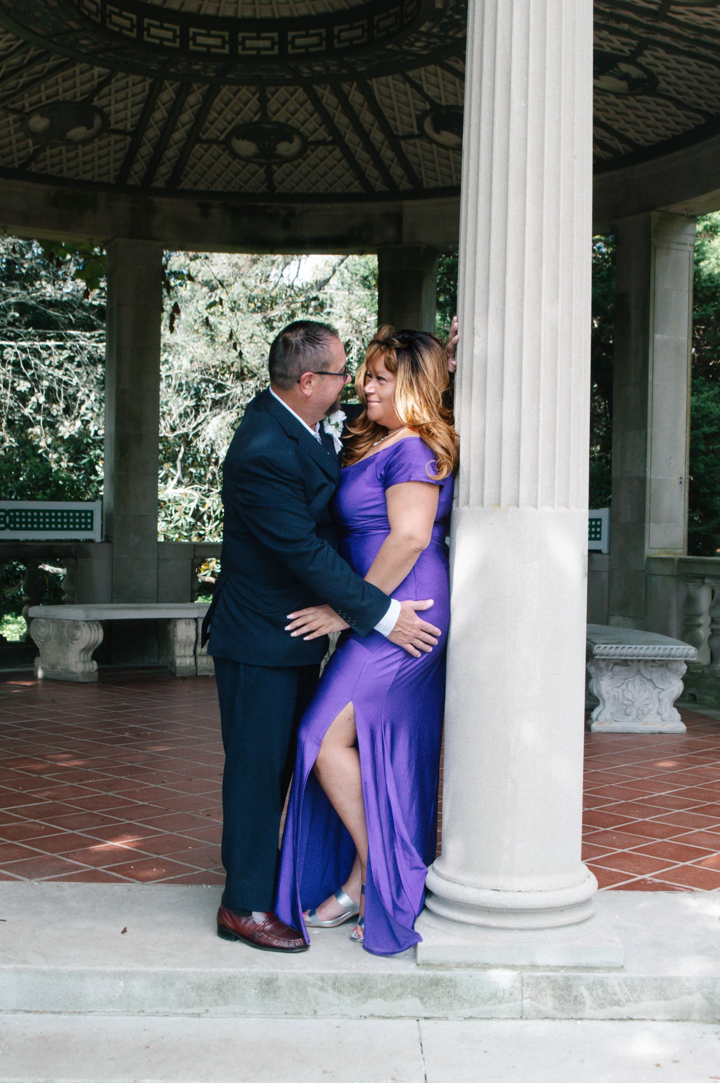 A couple standing close together under a gazebo, with the man in a black suit and the woman in a purple dress, gazing at each other affectionately.