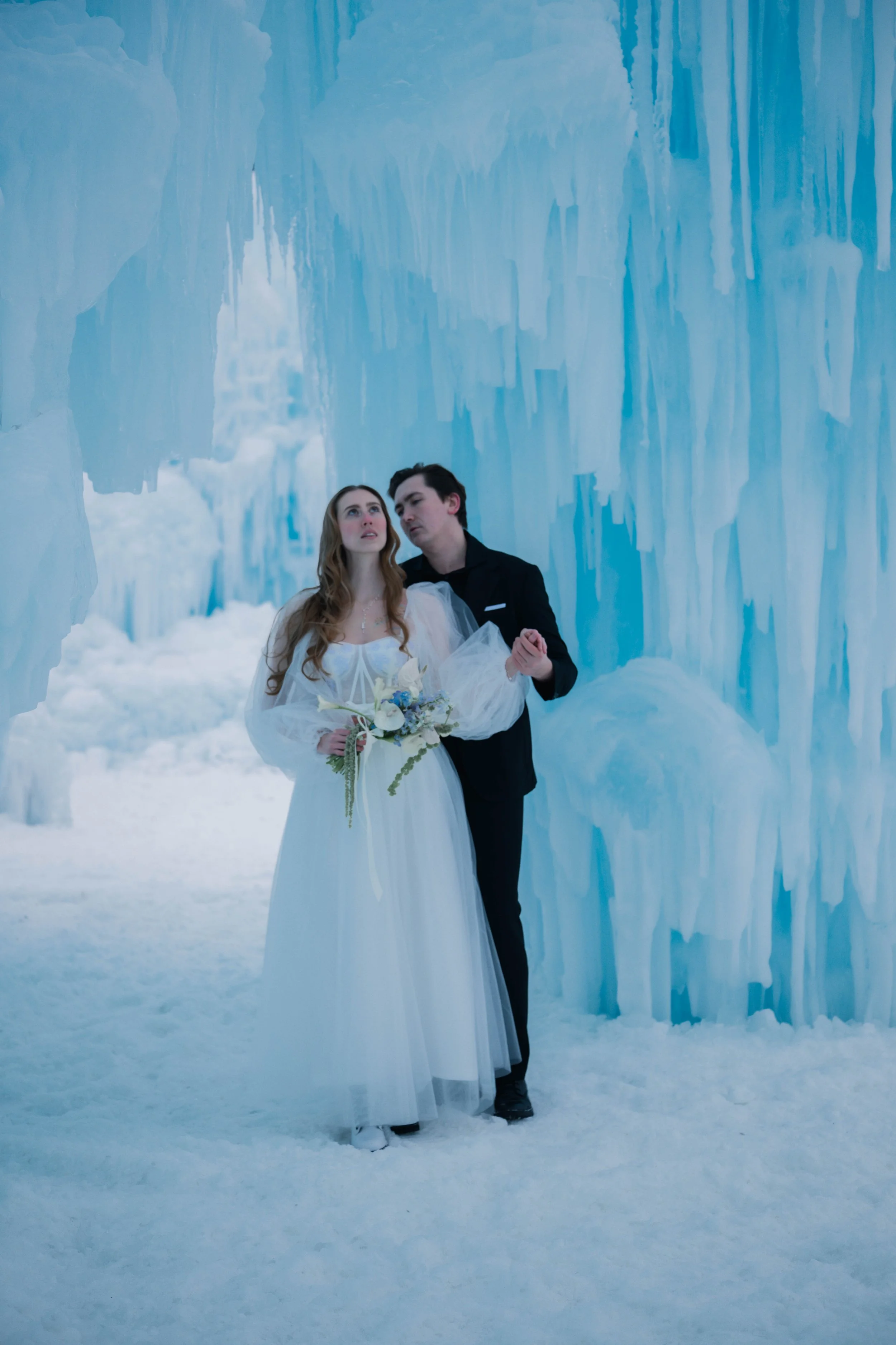 A bride in a white wedding dress and a groom in a black suit standing in an ice cave with blue ice formations.