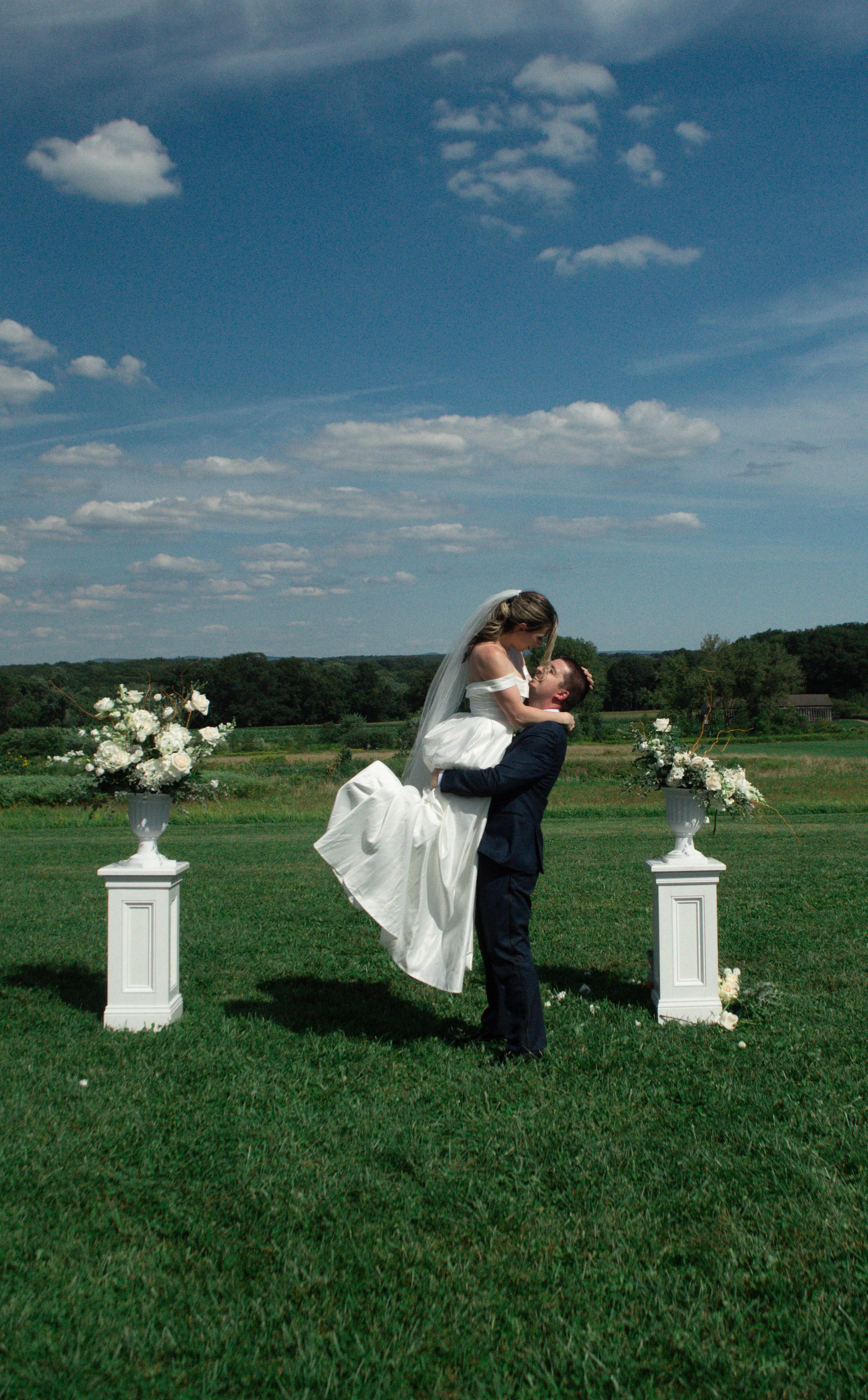 A newly married couple celebrating outdoors on a sunny day, with the woman in a white wedding dress and the man in a dark suit, holding her up in a field with floral arrangements on white pedestals.