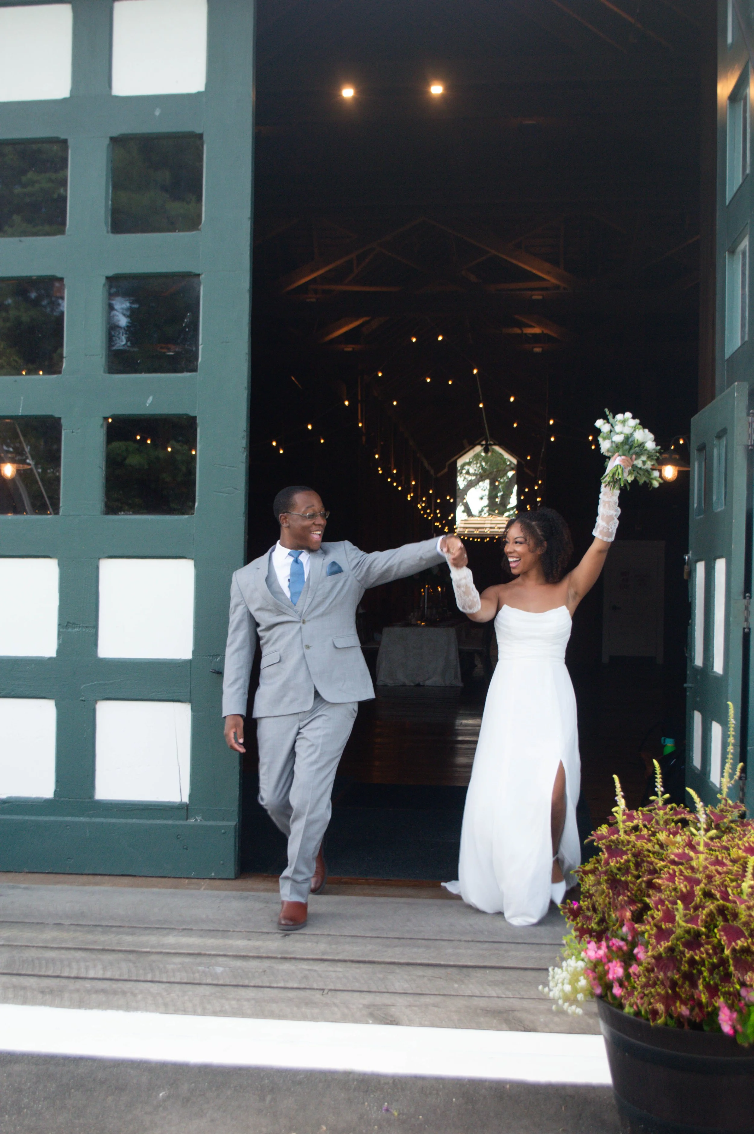 A newlywed couple exit a barn venue, smiling and celebrating. The bride in a white dress lifts a bouquet, while the groom in a gray suit holds her hand.