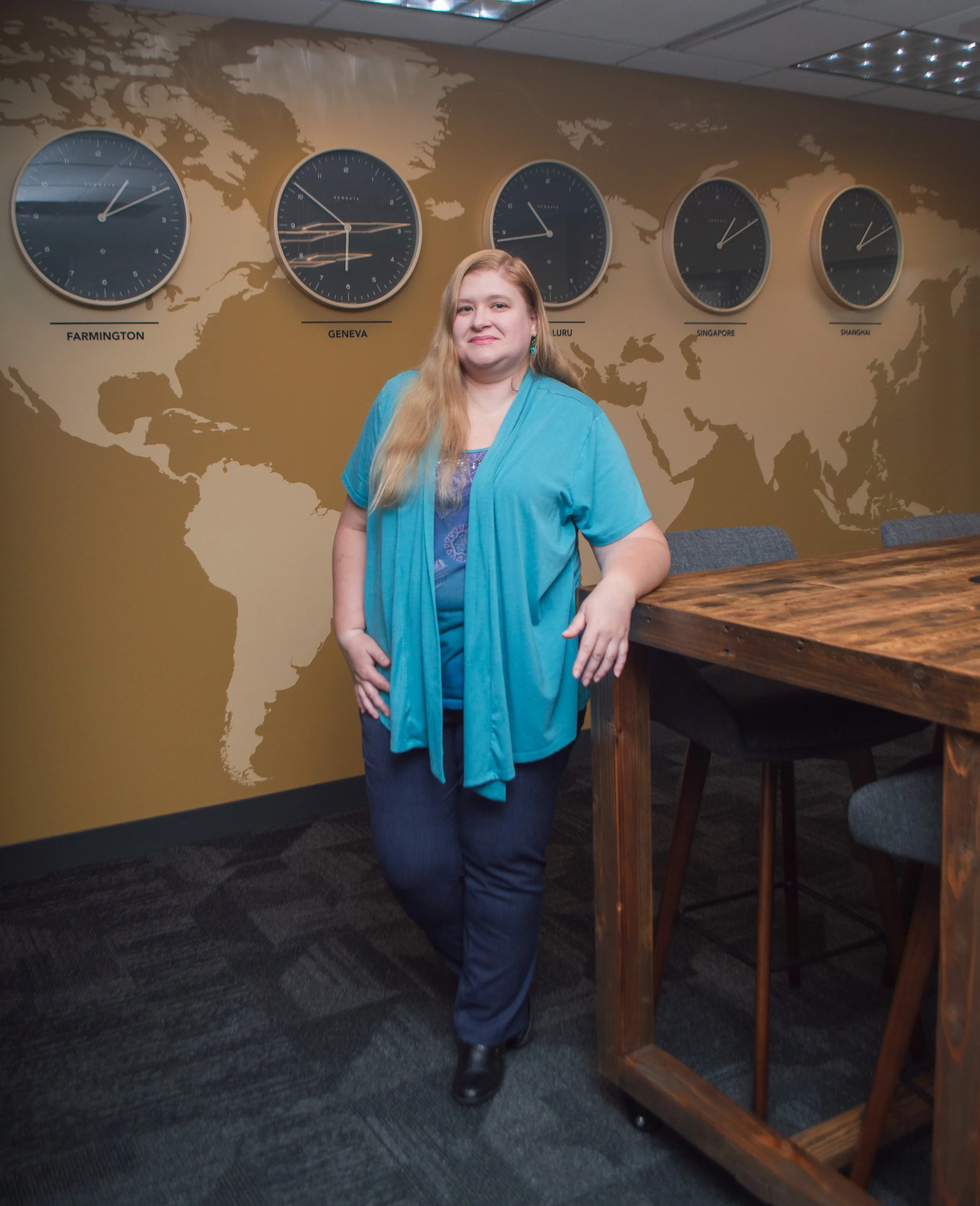 A woman with long blonde hair standing in a conference room next to a wooden table. Behind her on the wall is a world map with five large clocks showing different times labeled Farmington, Geneva, Bengaluru, Singapore, and Shanghai.
