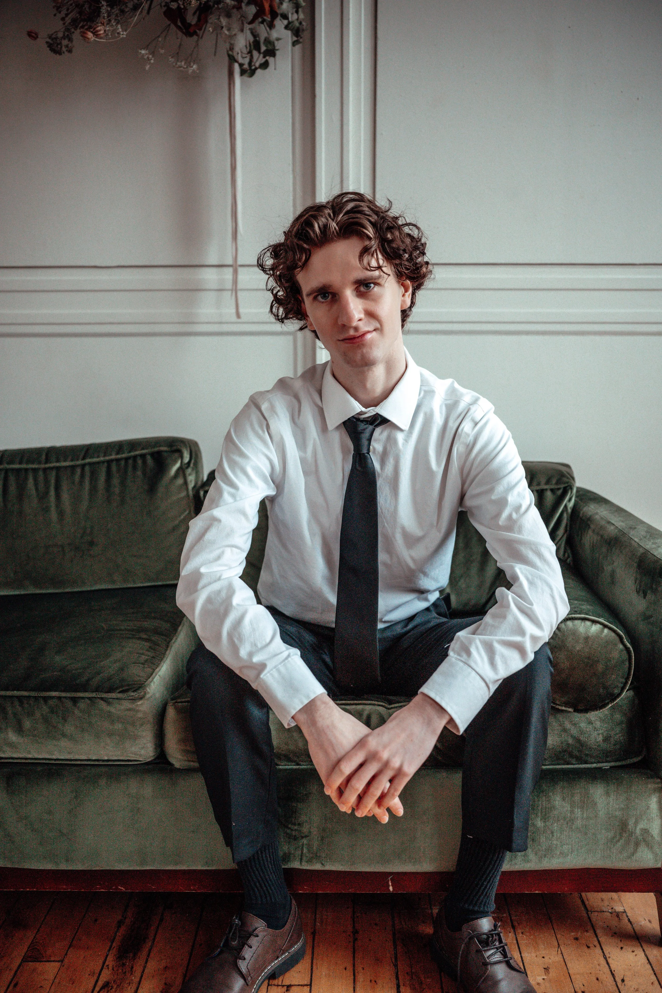 A young man with curly brown hair, wearing a white shirt, black tie, black pants, black socks, and brown shoes, sitting on a green vintage sofa in a room with light-colored walls and hardwood floor.