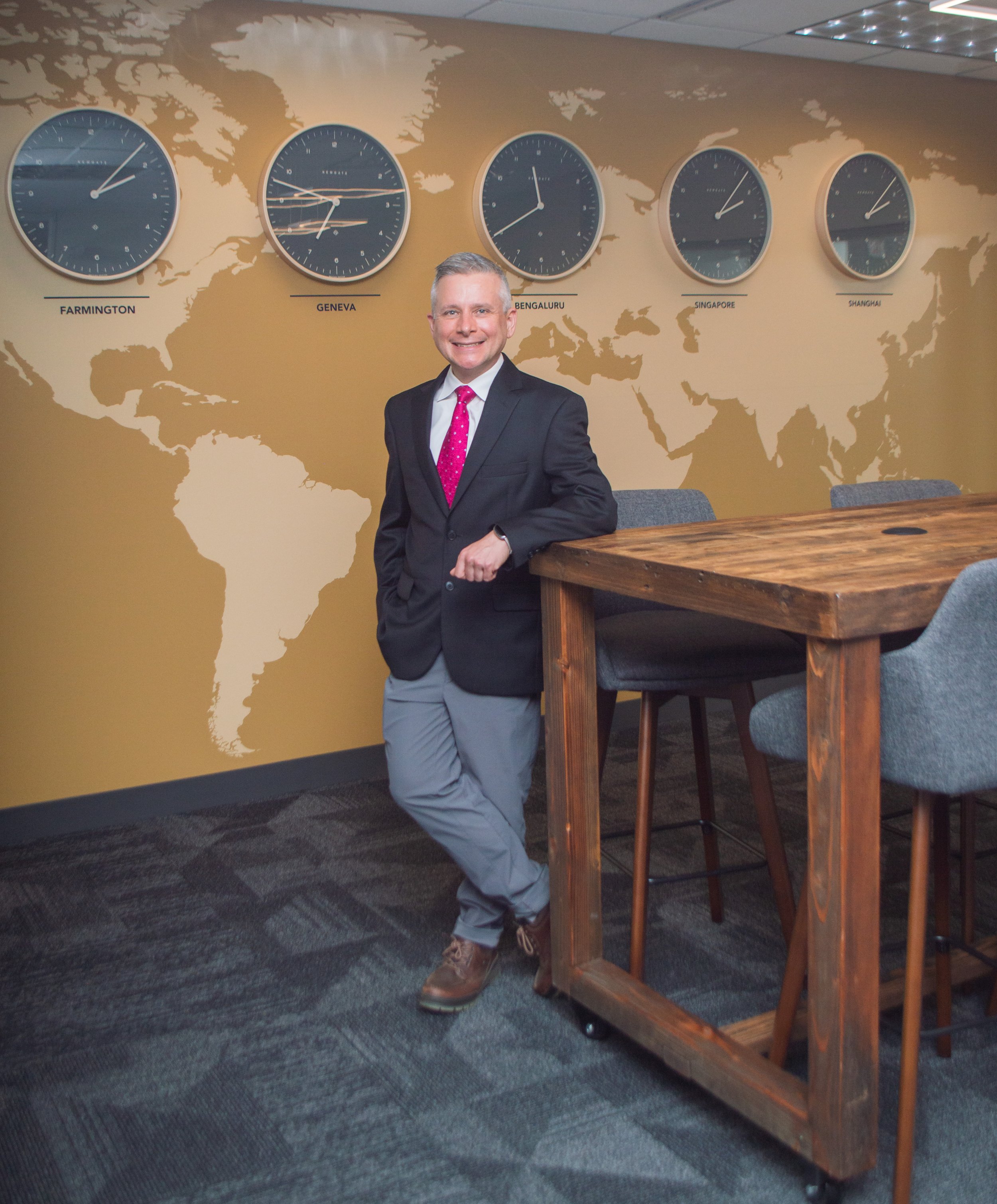 A man in a business suit standing in a conference room with a world map wall and five clocks showing different times labeled Farmington, Geneva, Bengaluru, Singapore, and Shanghai.