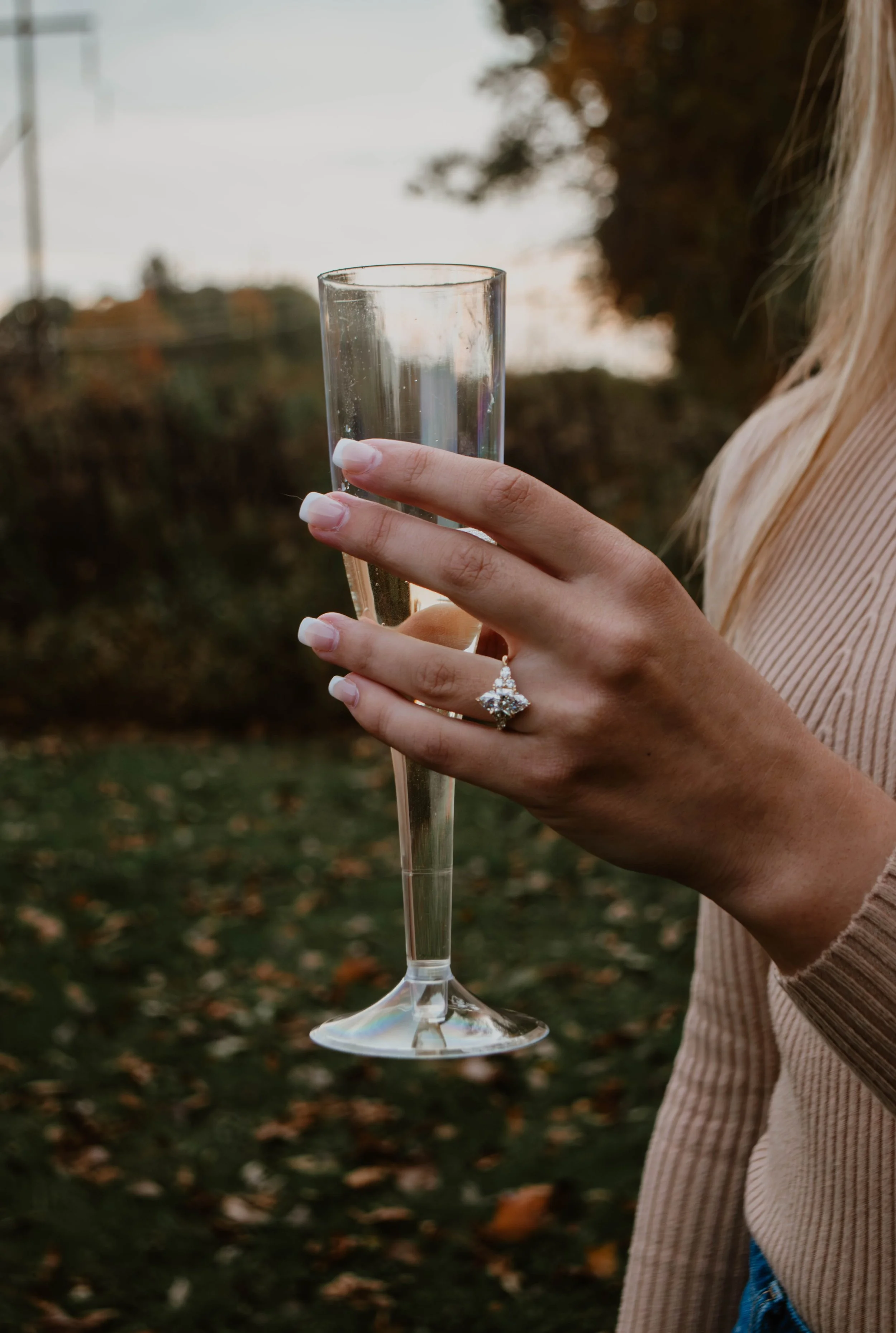A woman's hand holding a champagne flute with a sparkling ring on her finger, outdoors during fall.