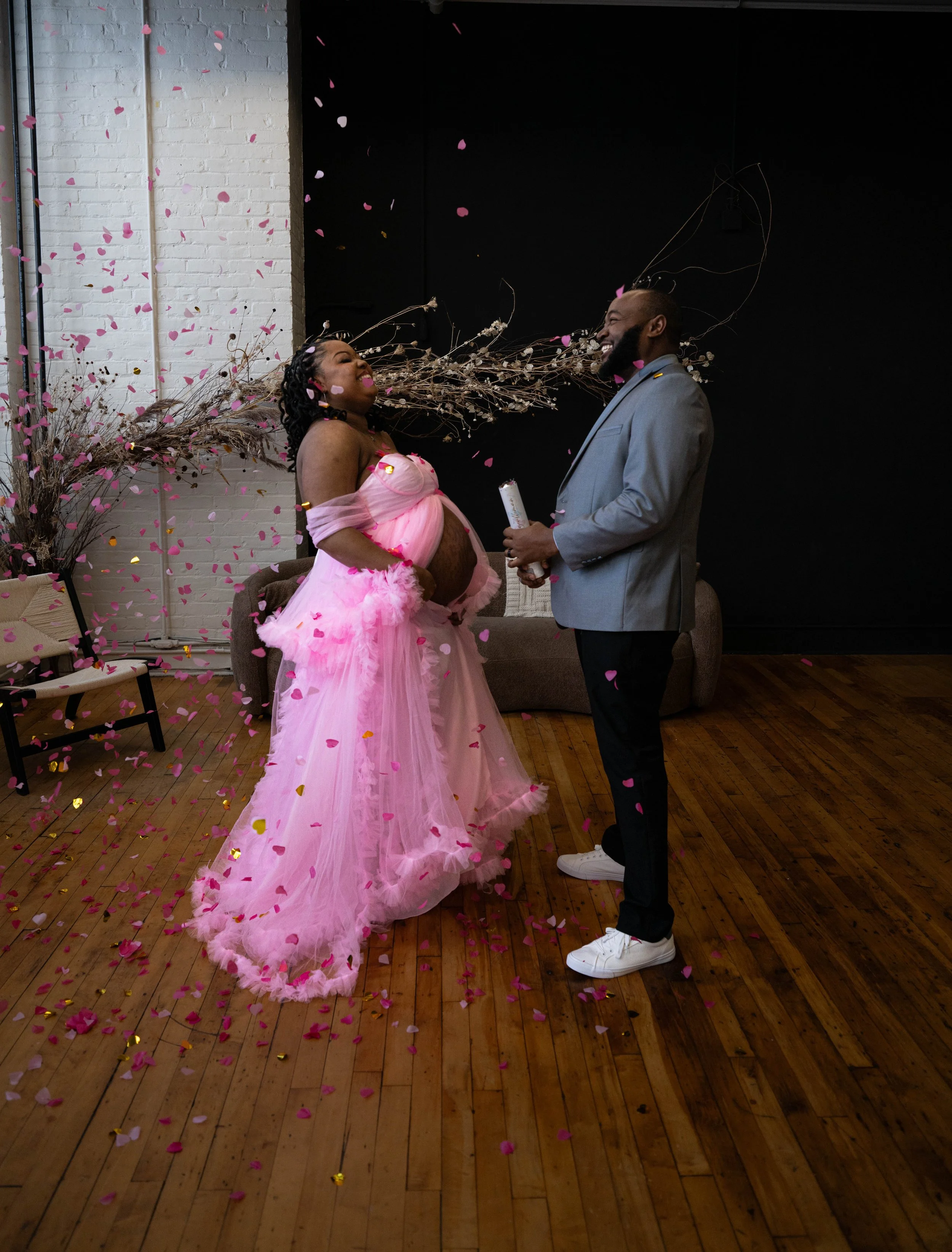 A pregnant woman in a pink dress celebrates a baby shower with a man, surrounded by pink and gold confetti and decorations, on a wooden floor with a white brick wall and dark background.