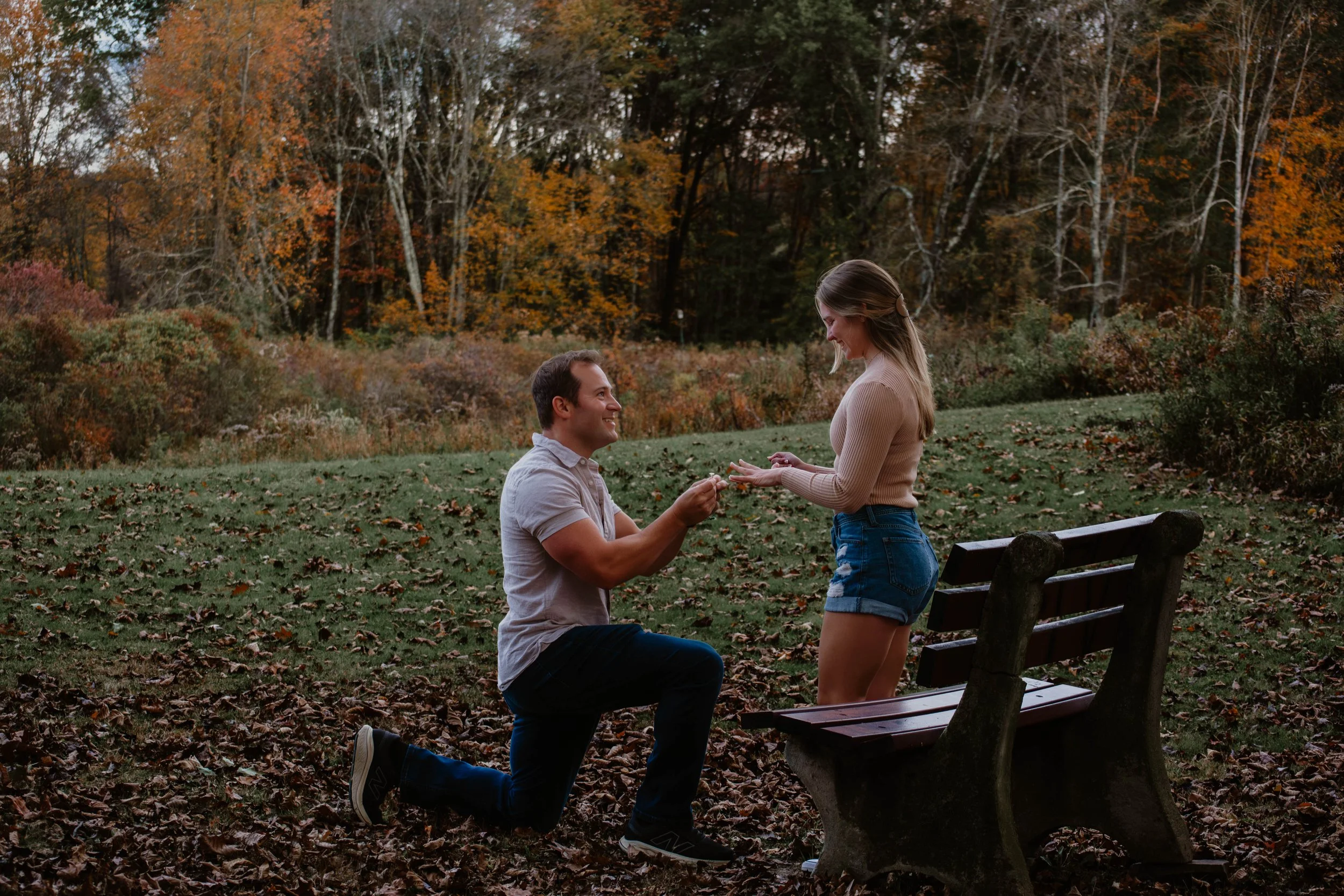 A man proposing to a woman in a park during autumn, with colorful fall foliage in the background.