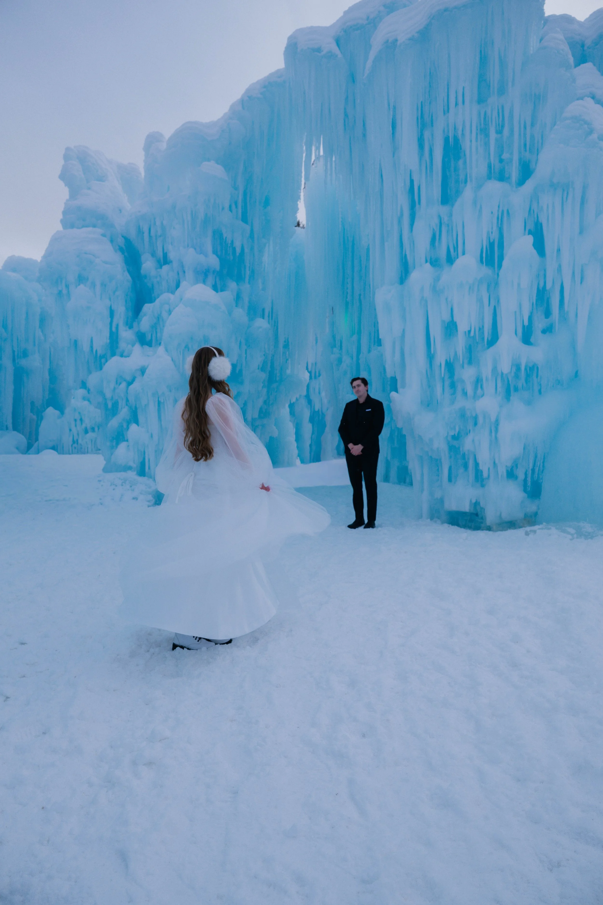 A woman in a white dress and a woman in a black suit stand in front of large ice formations in a snowy landscape.