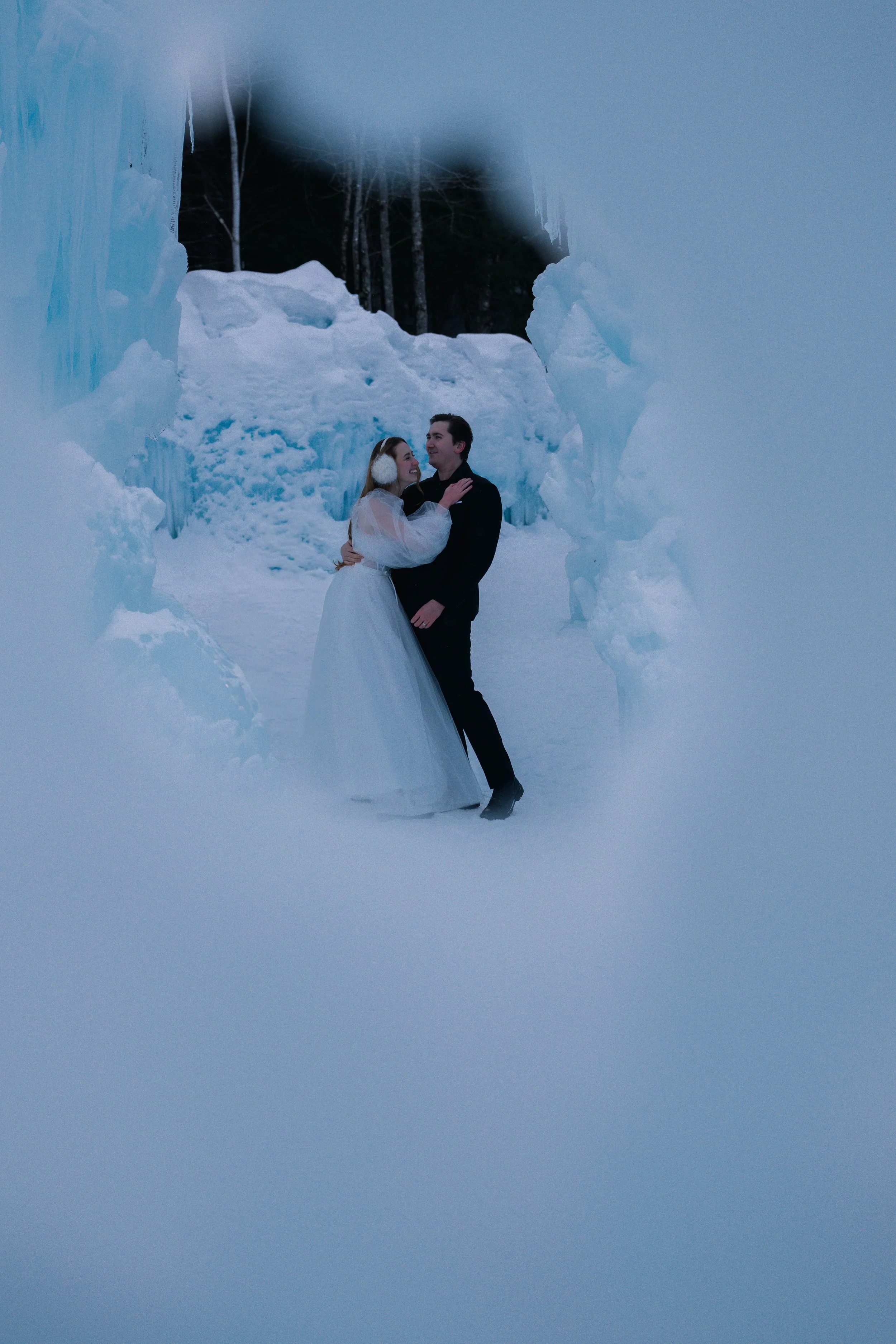A couple dressed in wedding attire standing close together inside a snow cave, surrounded by large ice formations and snow, with a dark background.