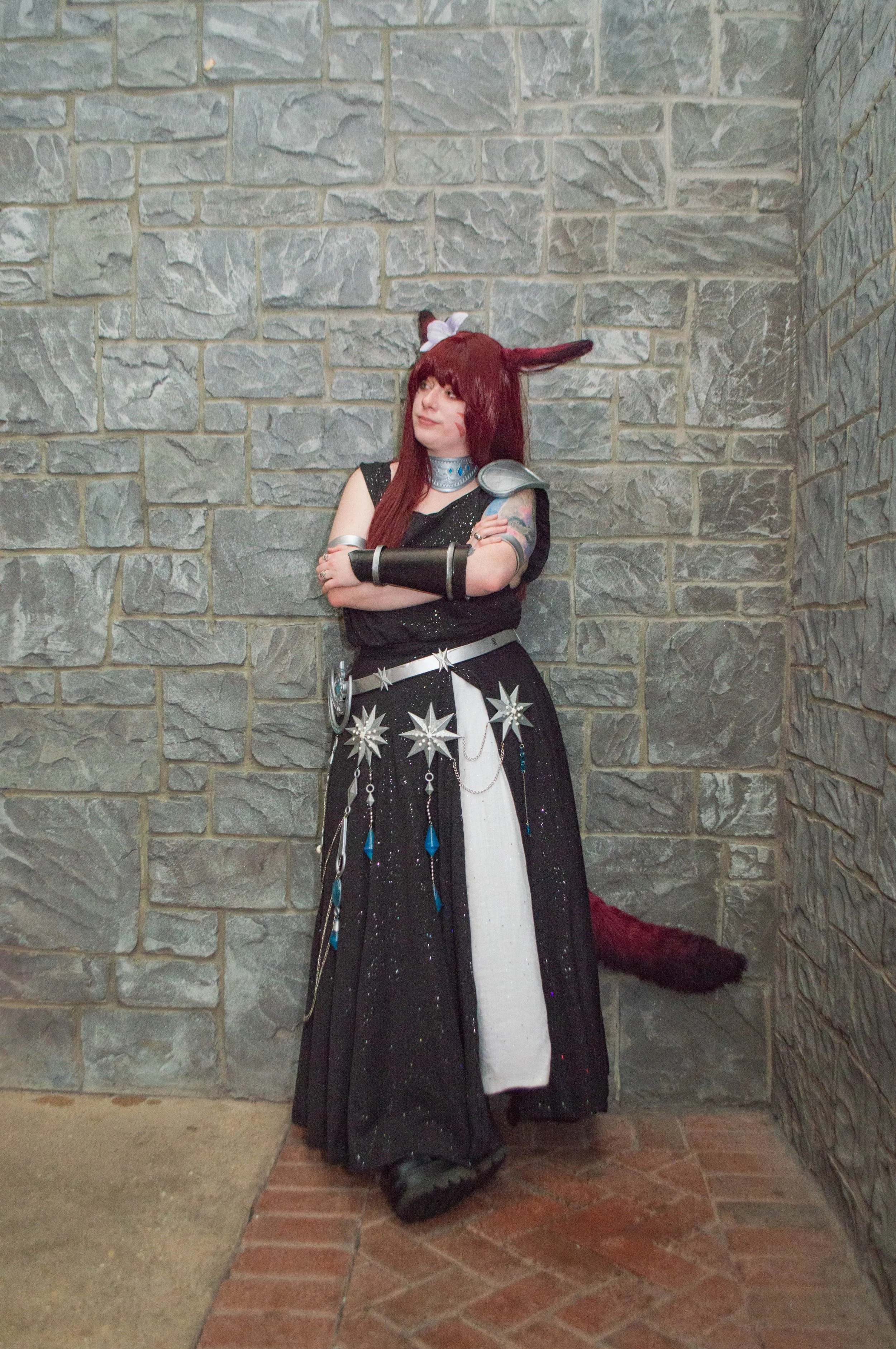 Person in a fantasy or cosplay costume with fox ears, a tail, and long red hair, standing against a stone wall with arms crossed.
