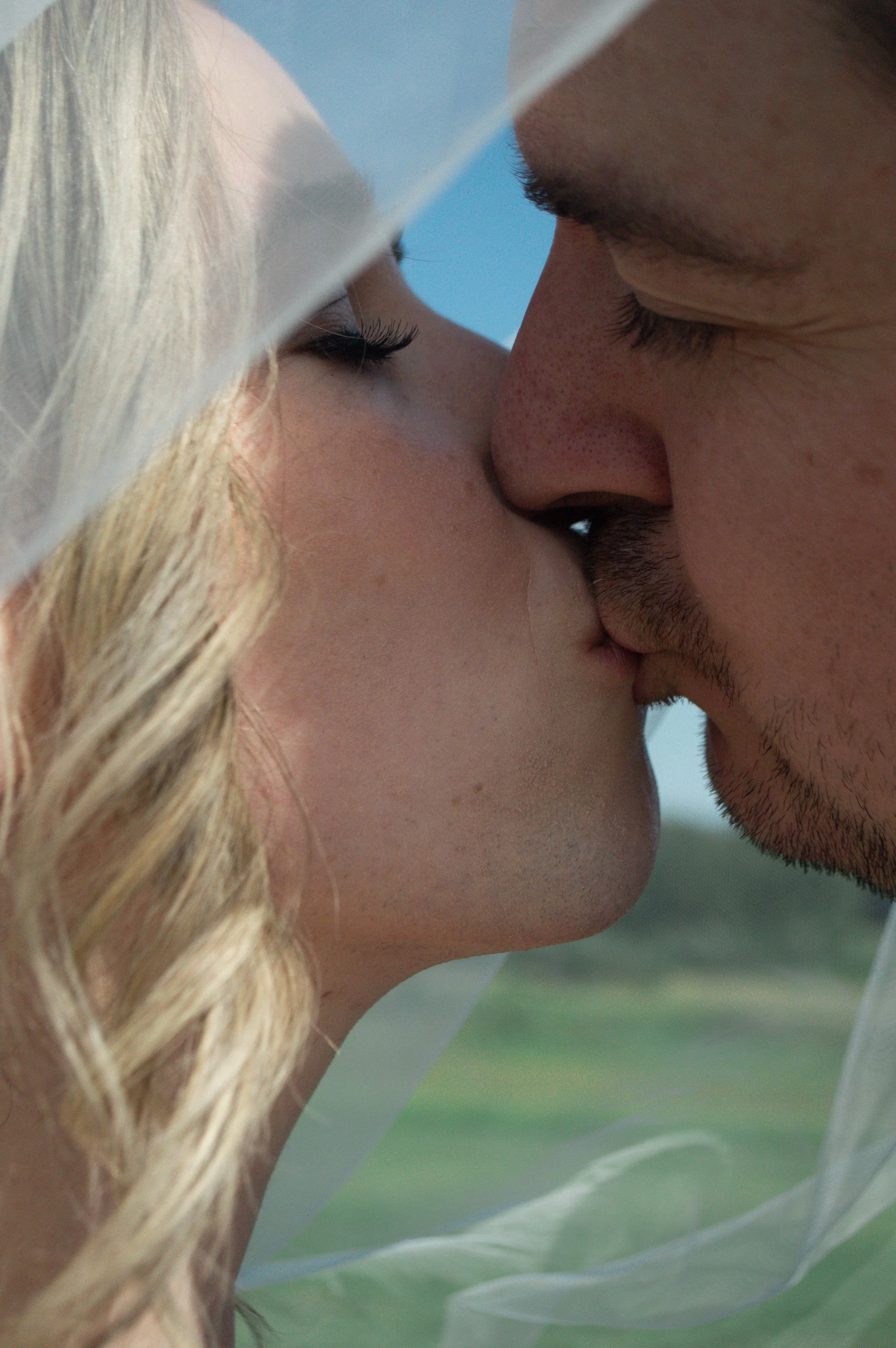 Close-up of a couple kissing, with a blurred green landscape and blue sky in the background.