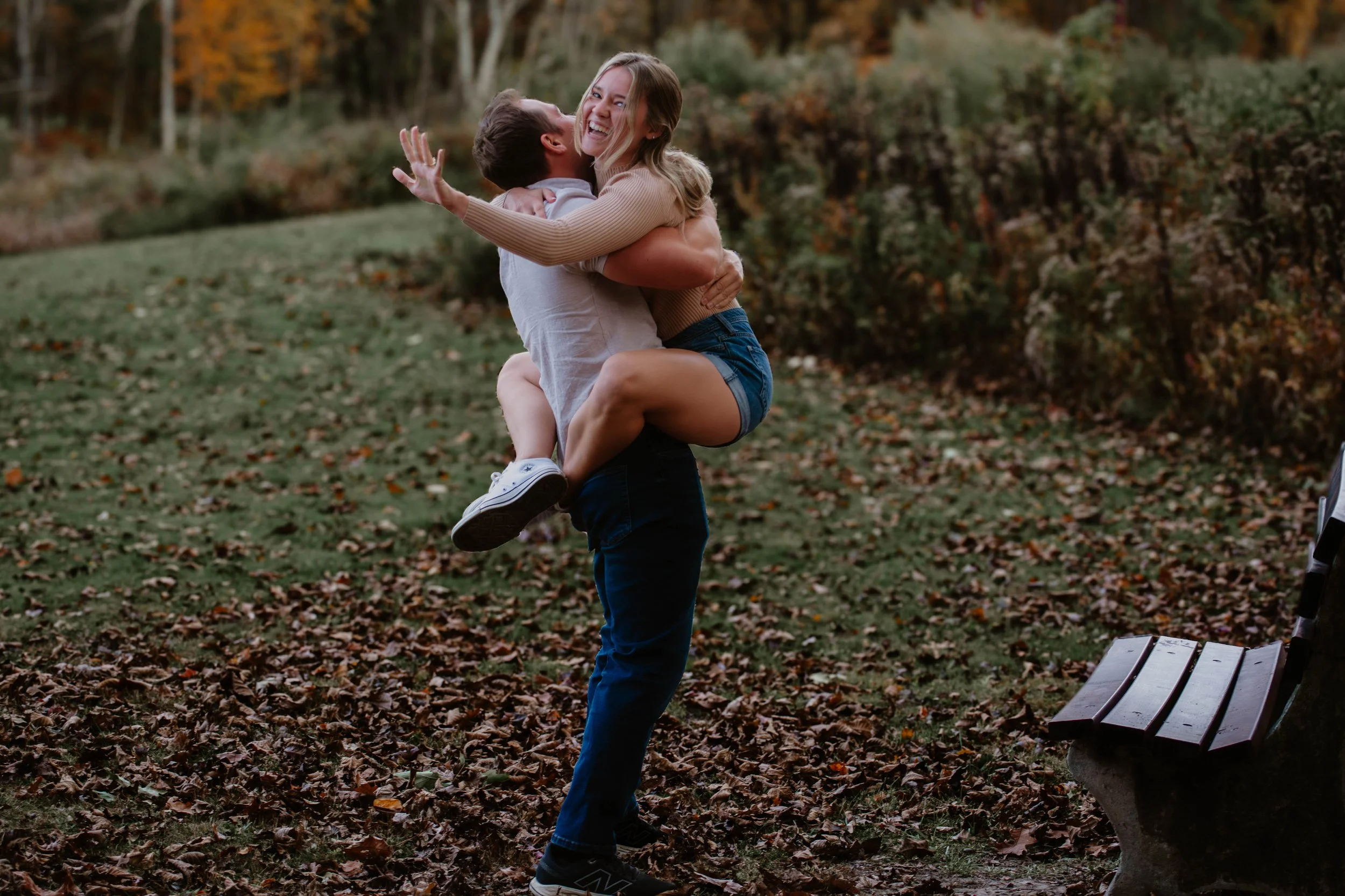A man lifting a woman in a park covered with fallen leaves during autumn, both smiling and embracing.