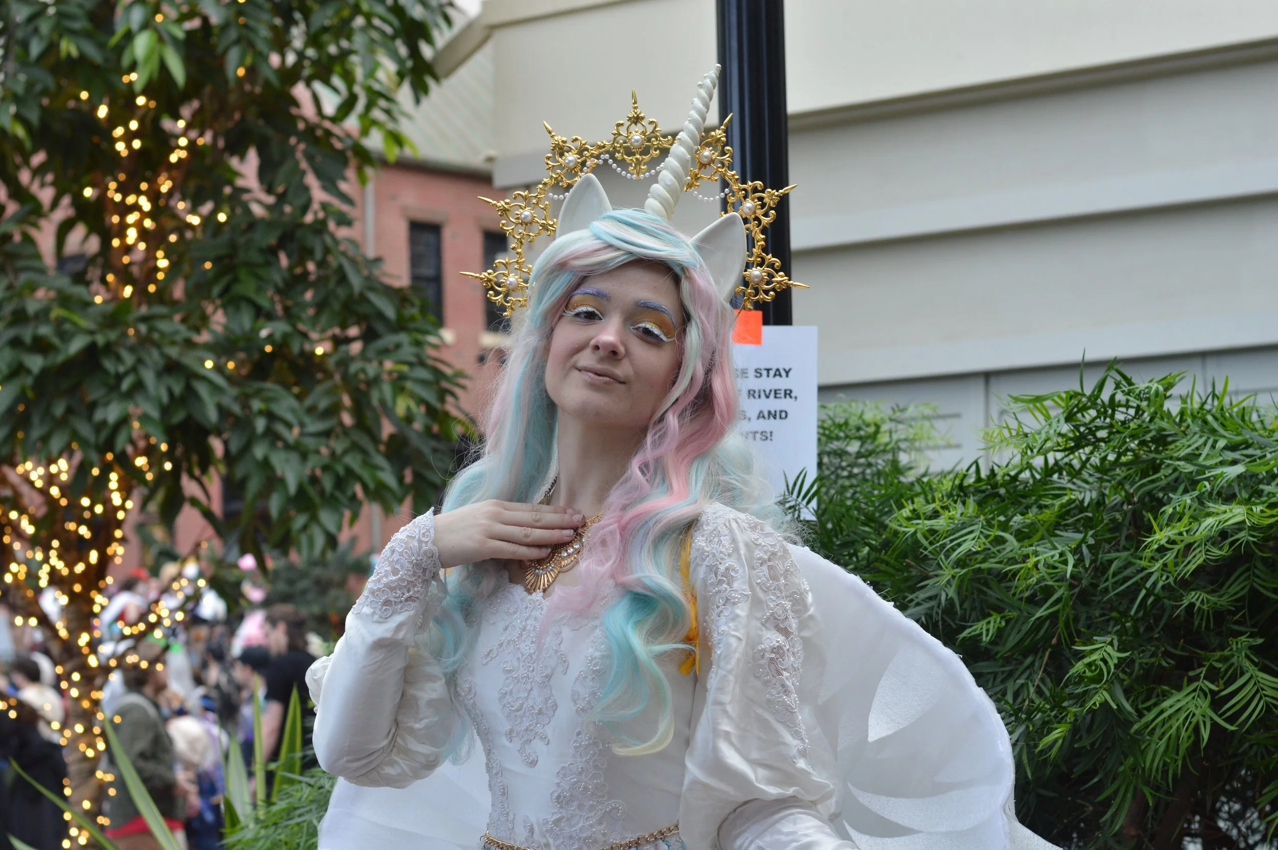 Person dressed as a unicorn with pastel-colored hair, a white dress with lace, gold jewelry, and a unicorn horn headpiece, standing outdoors among trees and buildings.