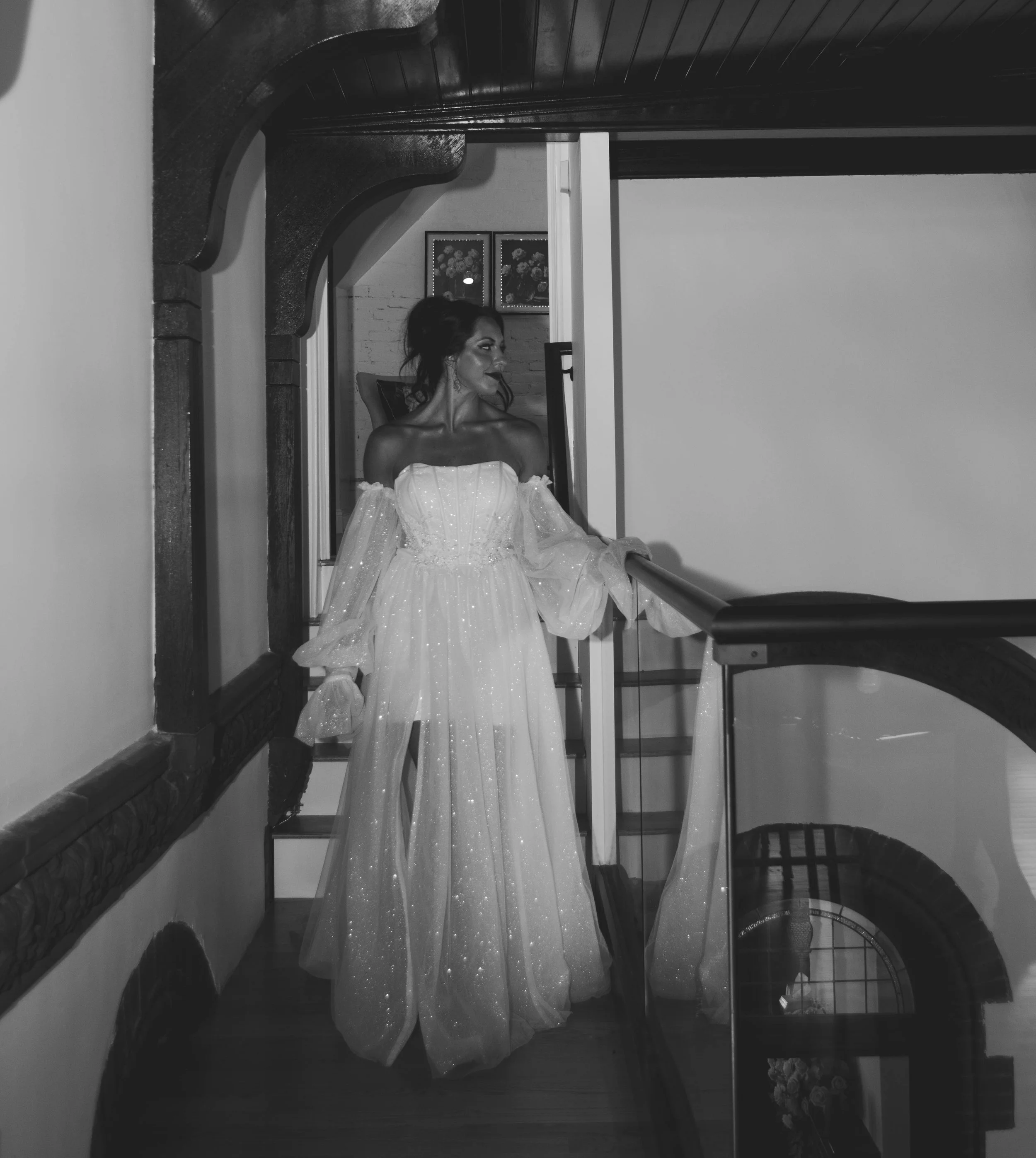 A woman in an elegant, off-the-shoulder, flowing gown with sparkles, standing on a staircase in a dimly lit interior, looking to the side.