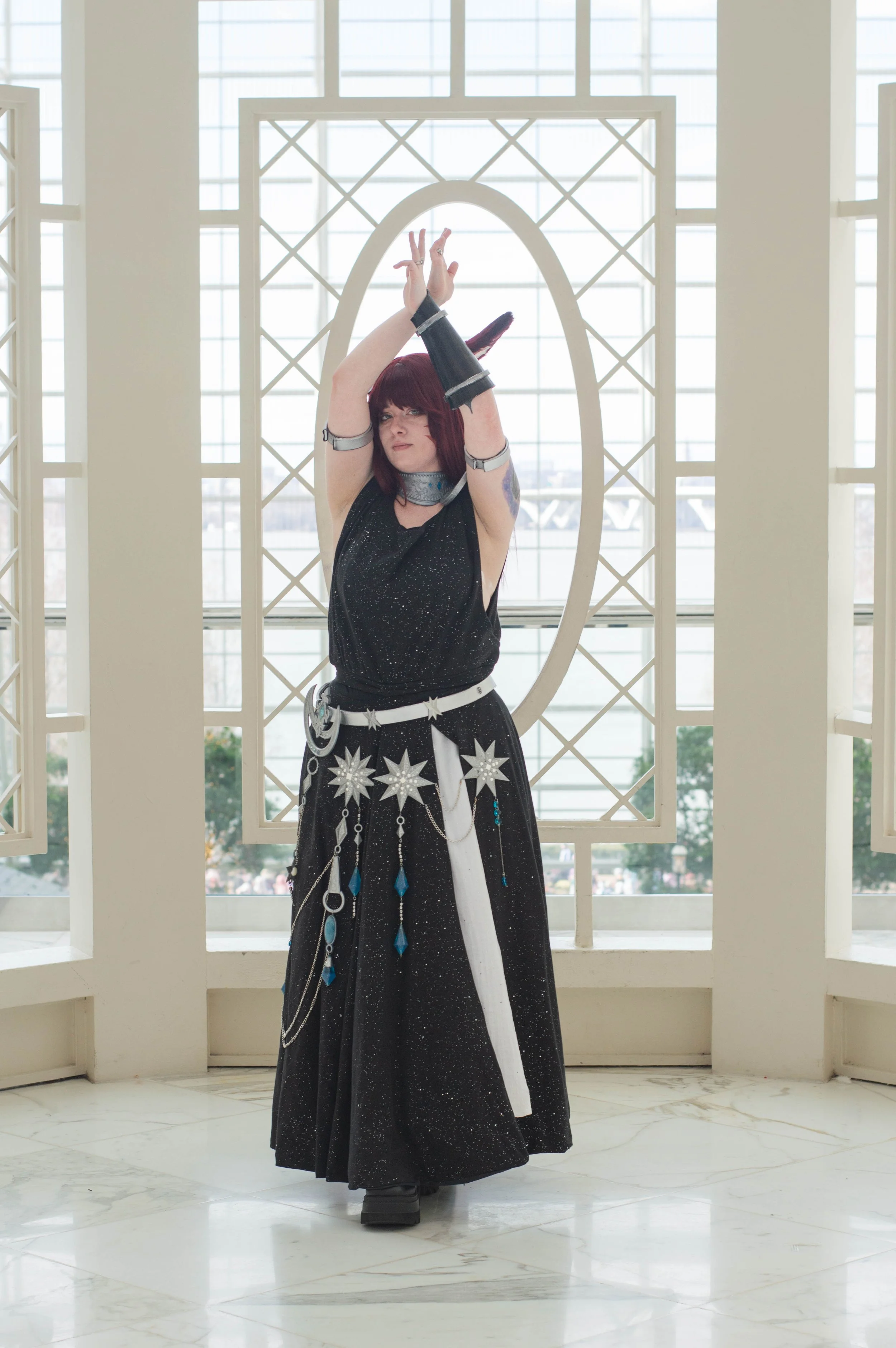 A person dressed in a black, starry-themed costume with metallic and blue accents, standing in front of a large window and decorative frame, striking a prayer-like pose with arms raised above their head.