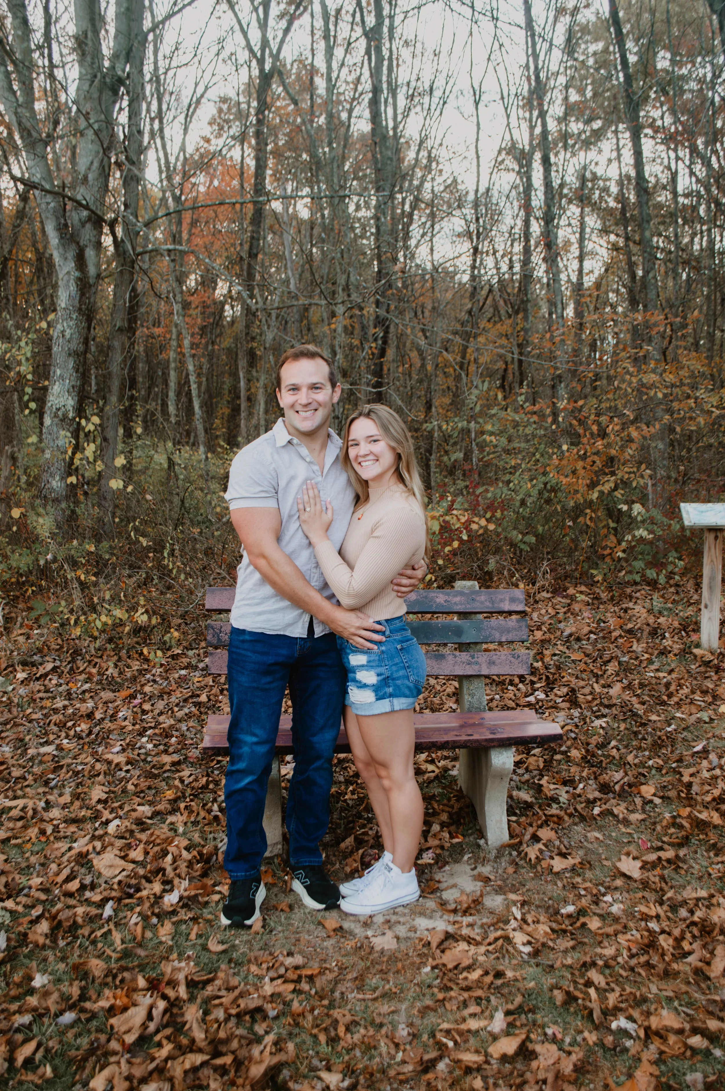 A smiling couple hugging in a park during fall, standing in front of a wooden bench surrounded by fallen leaves and trees with autumn foliage.