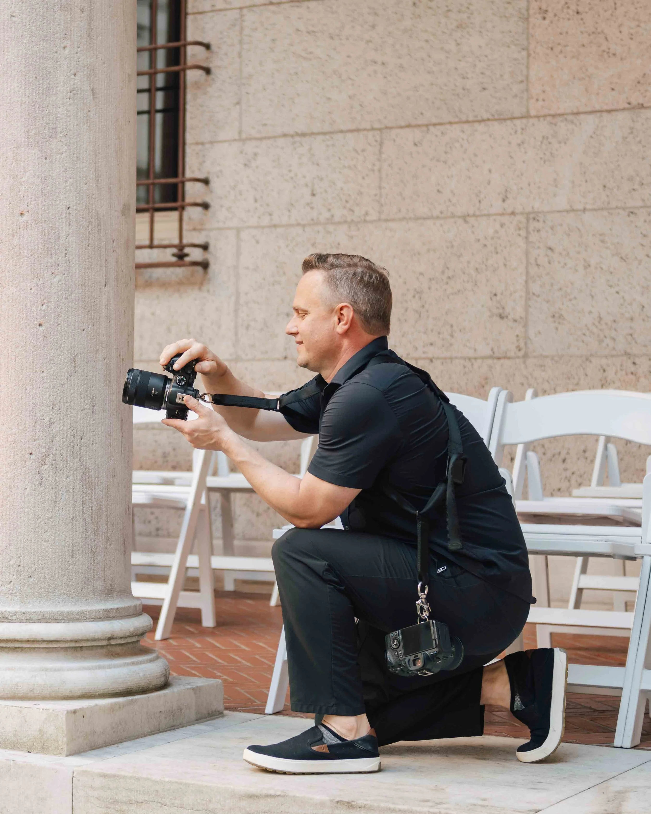 A man kneeling on one knee taking a photograph with a DSLR camera, wearing black clothing and sneakers, with additional camera gear hanging from his neck.