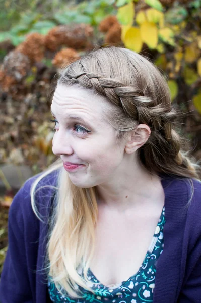 A white woman smiling off camera. The picture is a her profile and you can see fall foliage behind her. Her blonde hair is braided in a large dutch braid that is predominant in the shot.
