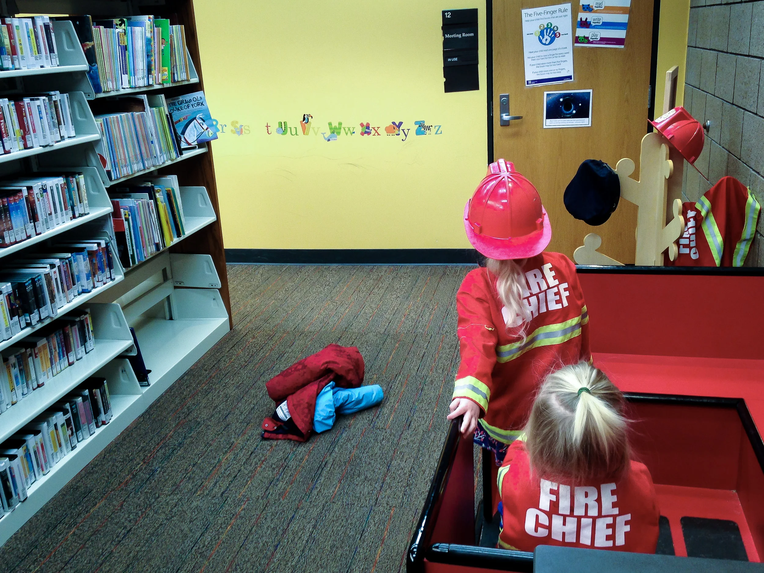 Image: Two blonde children with dress up fire chief coats and hats looking at a yellow wall with alphabet stickers. Their red and blue coats are thrown on the floor next to a very large aisle shelf of books and audio books.