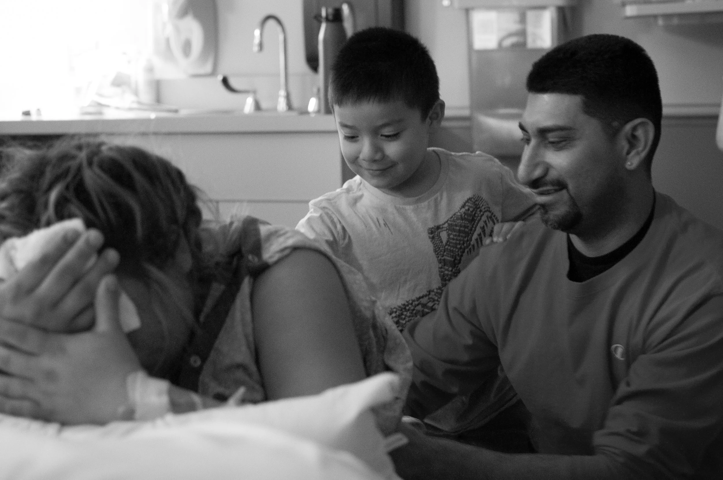 A birthing person is leaning over with their face buried in their hands. Behind them is a a elementary aged child rubbing their back next to their support person who is also rubbing the birthing person’s back.