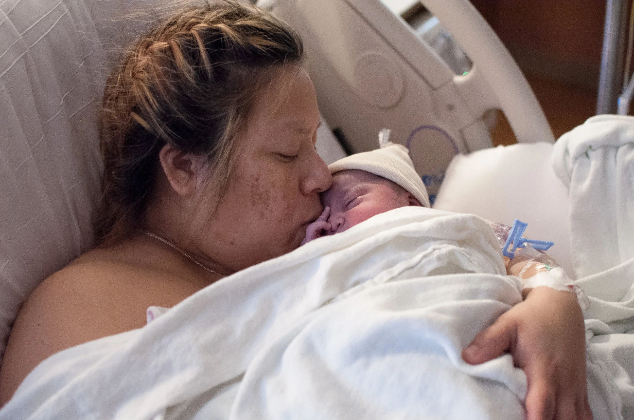 A birthing person holding their newborn close and kissing them on the face. The birthing person has french braids and is laying skin to skin with baby under a sheet.