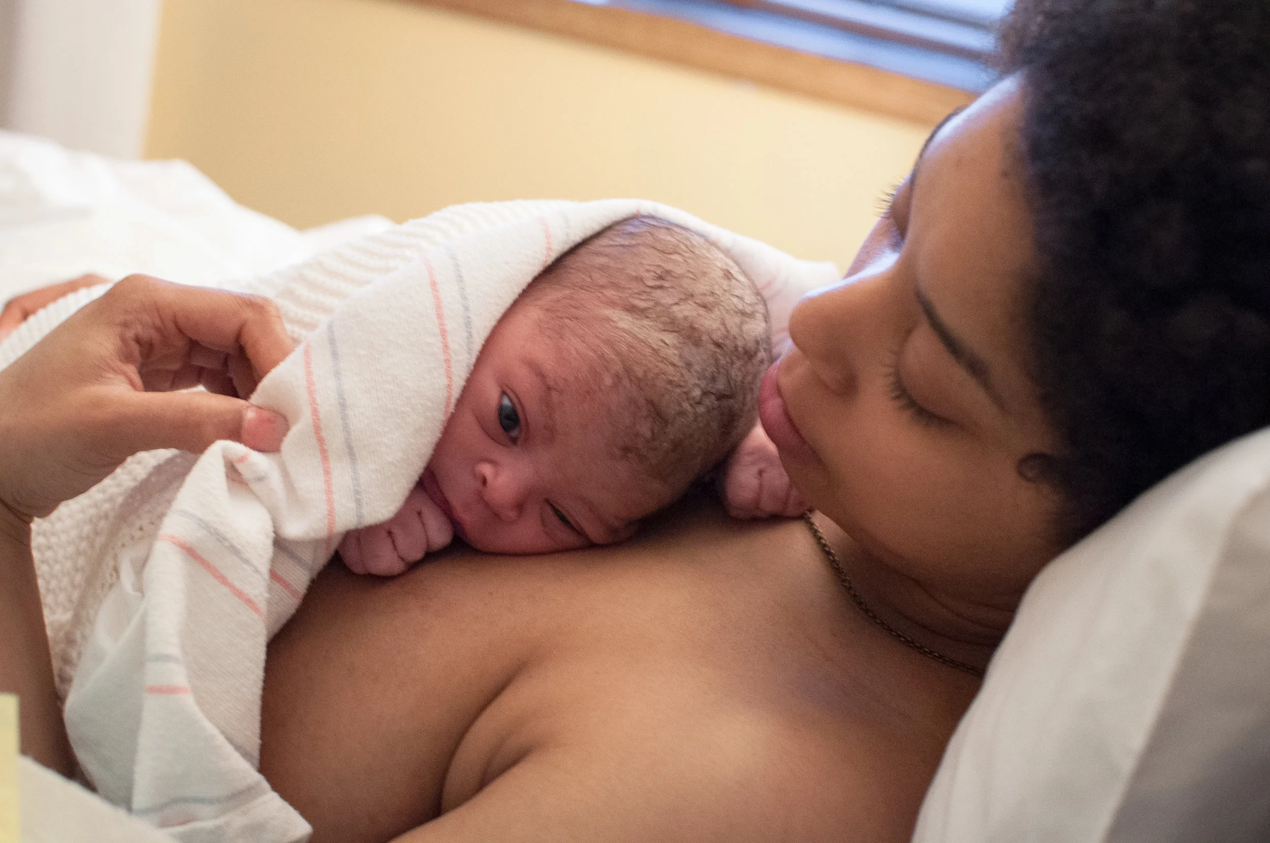 A birthing person laying bare chested on a bed. There is a newborn under a blanket with their hand to their mouth and looking off camera. The birthing person is looking down lovingly and is adjusting the blanket with their hand.