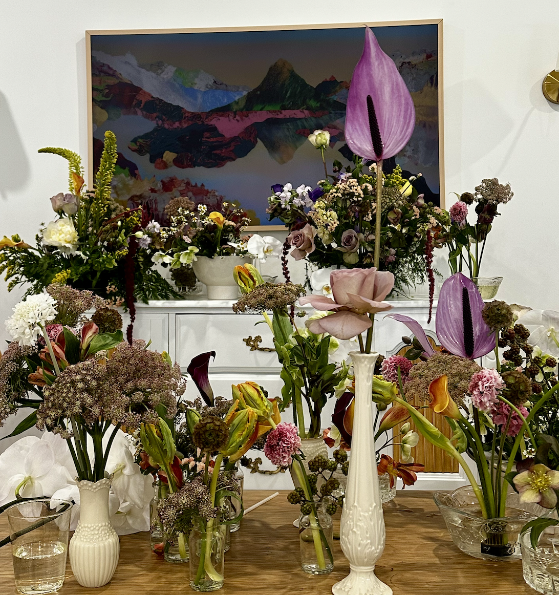 A room filled with numerous glass vases and vases of different shapes, holding a variety of fresh flowers including roses, calla lilies, anthuriums, and other colorful blooms placed on a wooden table. In the background, there's a white cabinet and a 