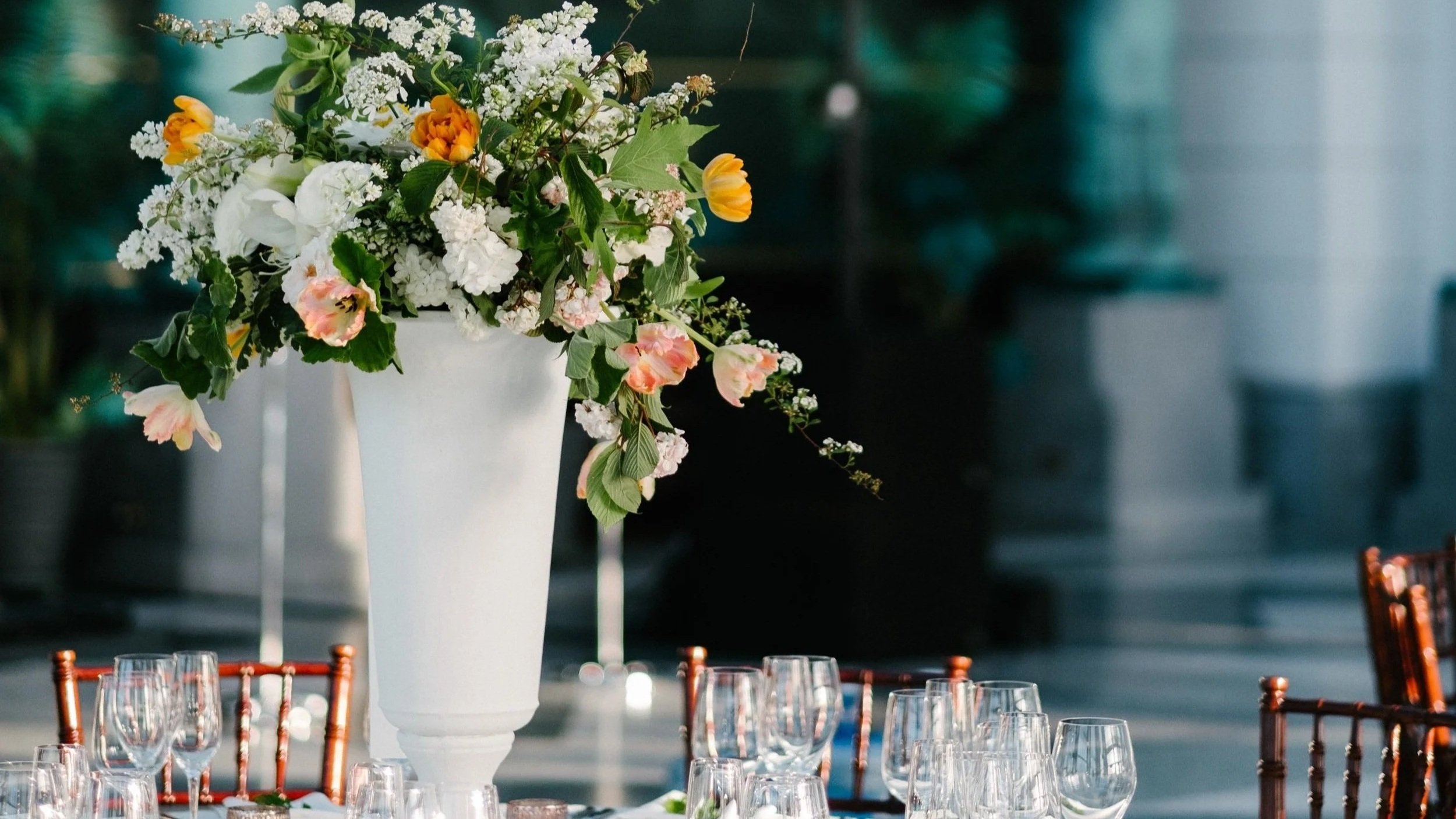 Elegant table setting with a large floral centerpiece in a white vase, surrounded by glassware and candles, at an outdoor event with a green tent in the background.