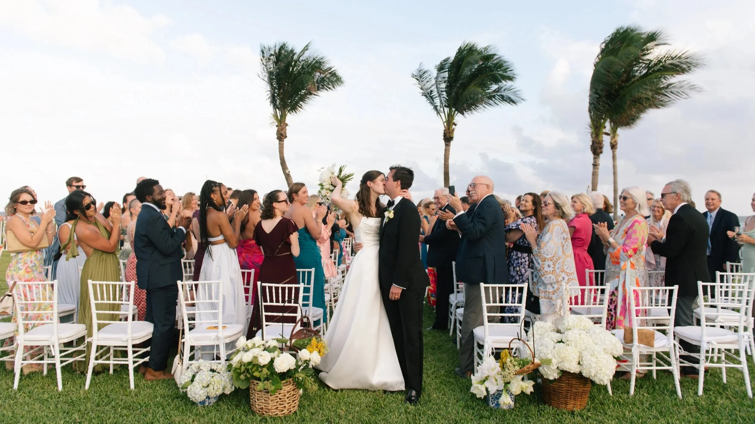 A wedding ceremony outdoors with a bride and groom kissing, surrounded by guests clapping and celebrating, on a grassy area with palm trees and a partly cloudy sky in the background.
