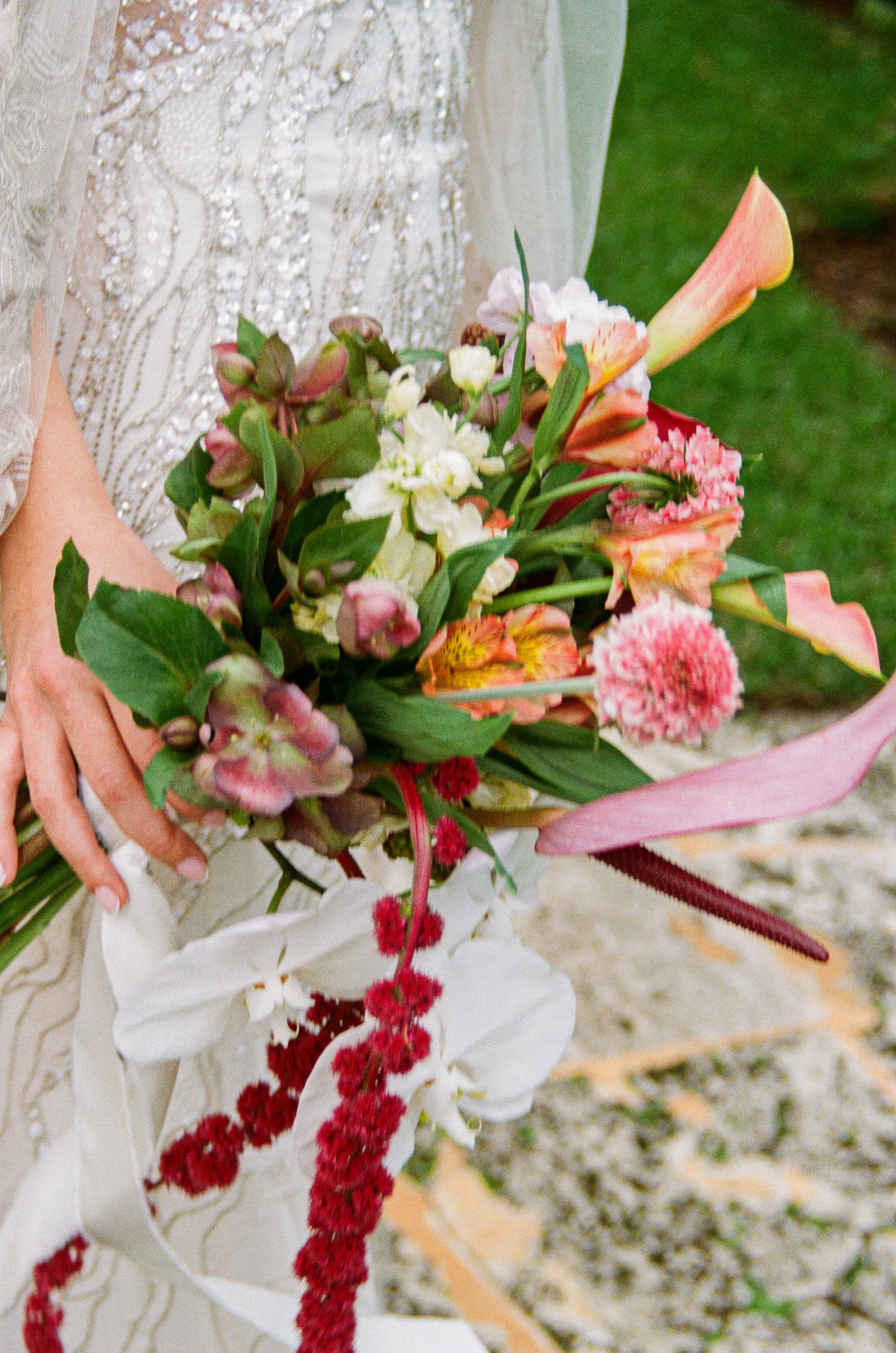 A person wearing a wedding dress holding a bouquet of mixed flowers, including lilies, calla lilies, and other colorful blooms, with a blurred outdoor background.