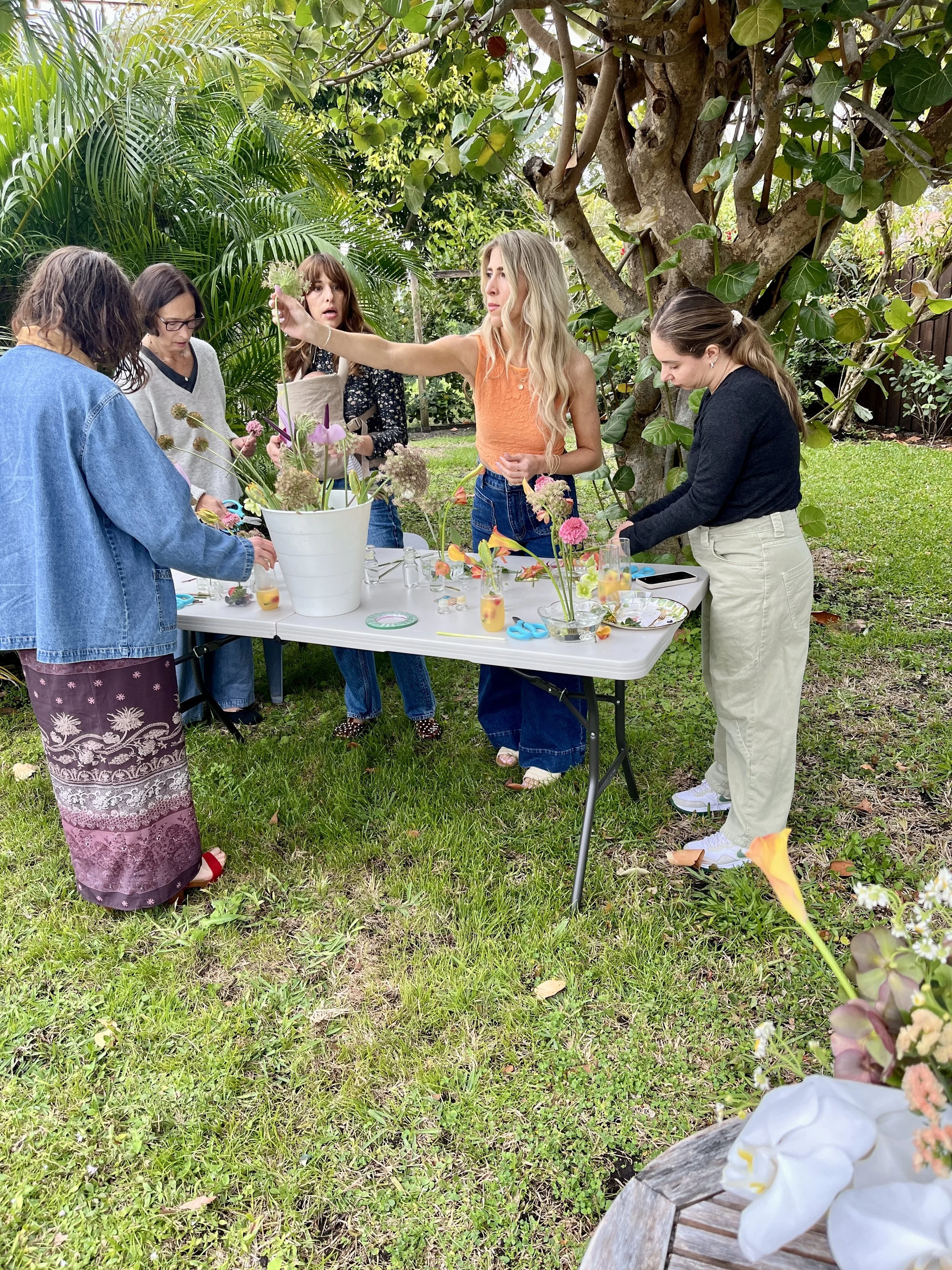 Group of women gathered around a table outdoors, arranging flowers in a garden setting with greenery and trees.