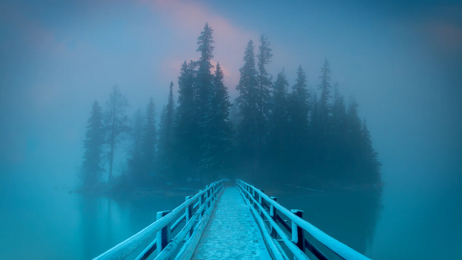 A snowy dock extending over a calm lake, with misty evergreen trees and mountains in the background during dawn or dusk.