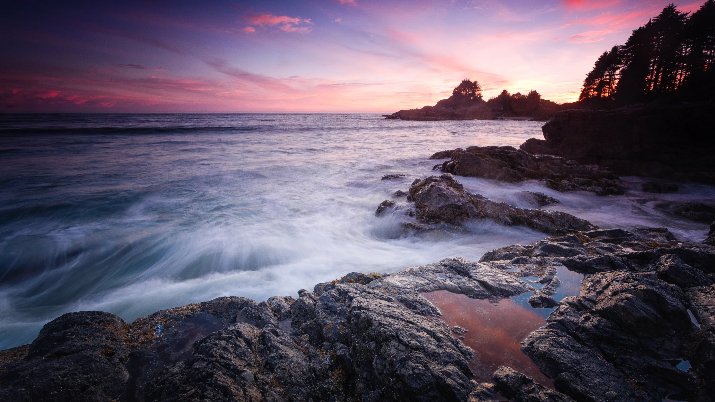 Sunset over a rocky shoreline with gentle waves and a tree-covered cliff in the distance.