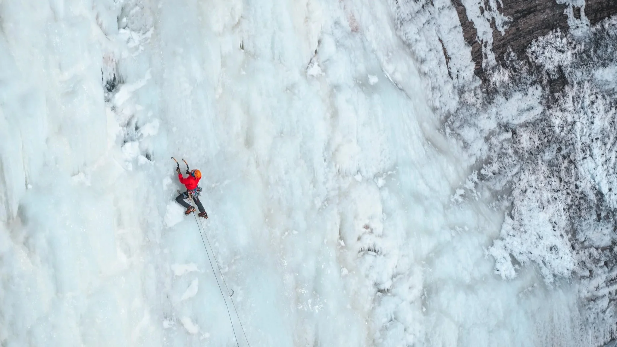 A person ice climbing on a frozen, icy wall with a harness and helmet, surrounded by snow and ice-covered rocks.
