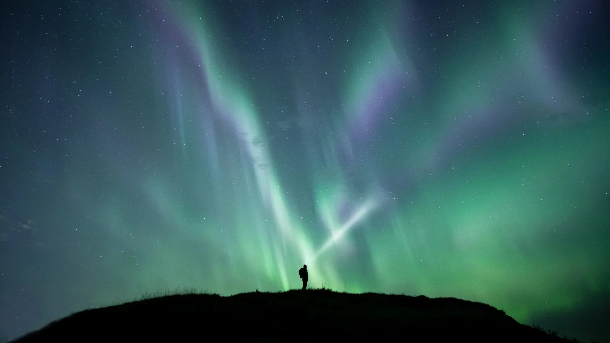 Person standing on a hill under the night sky with colorful northern lights and stars.