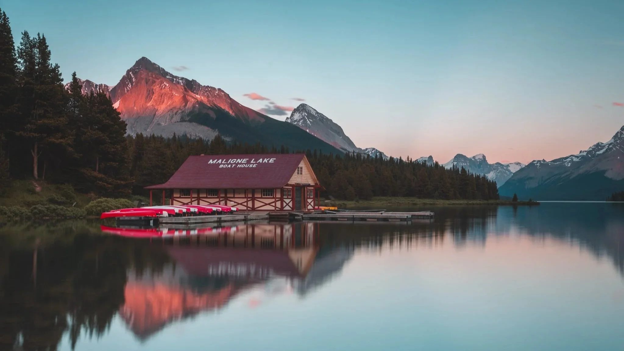 A wooden boathouse labeled 'Maligne Lake Boat House' situated by a calm lake with red boats docked, surrounded by forest, and mountains in the background during sunset.