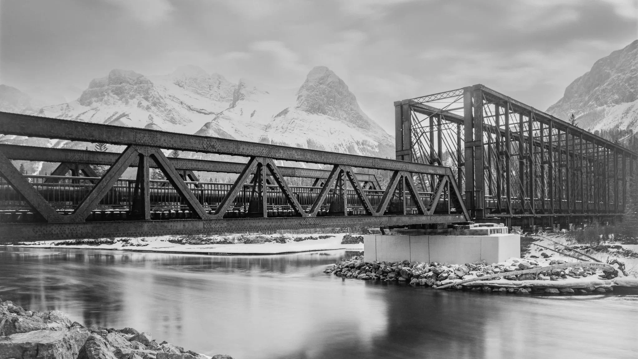 A black-and-white photo of a metal truss bridge crossing a river with snow-covered mountains in the background.