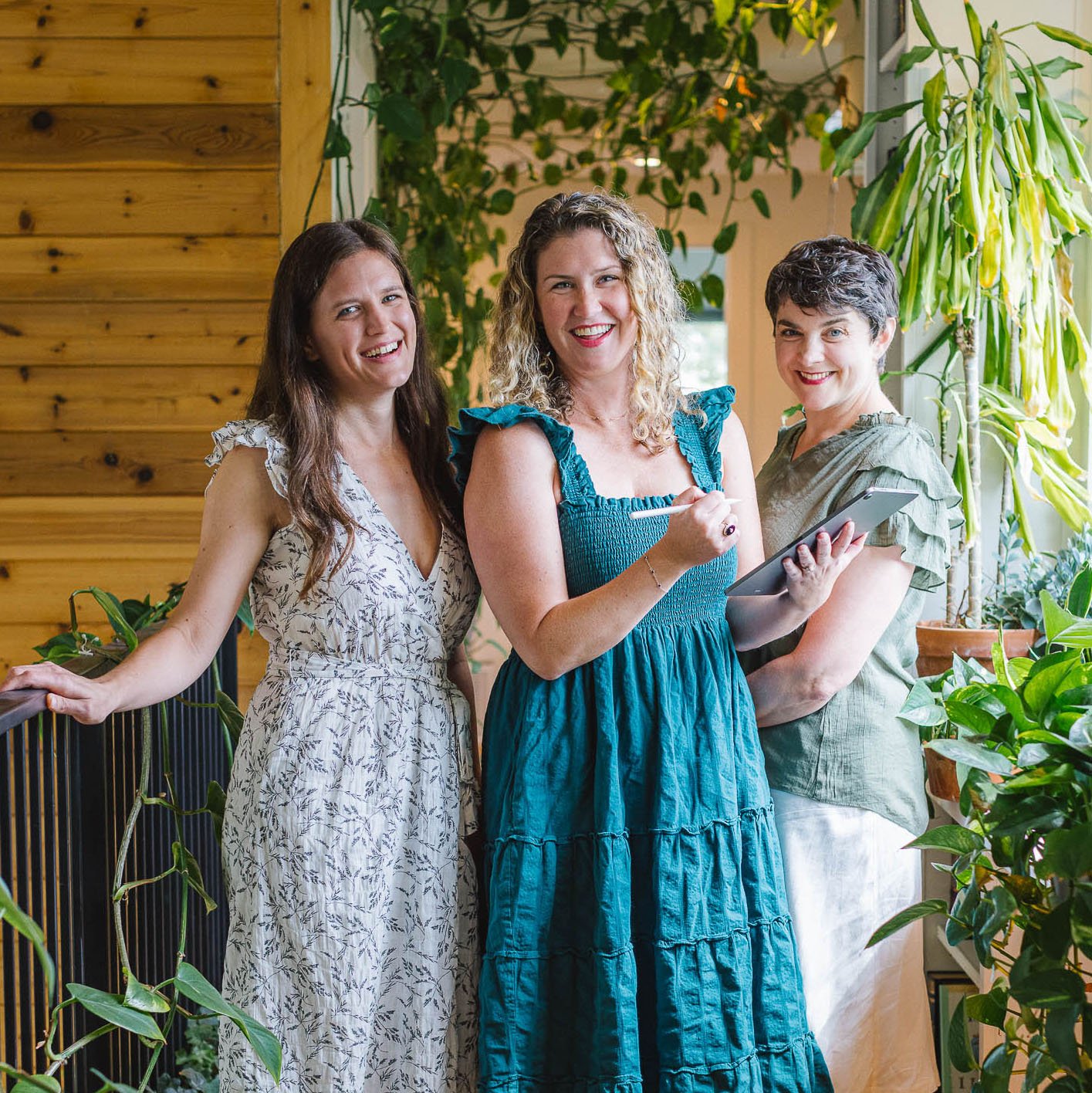Three-person business team smiling together in a bright, plant-filled workspace.