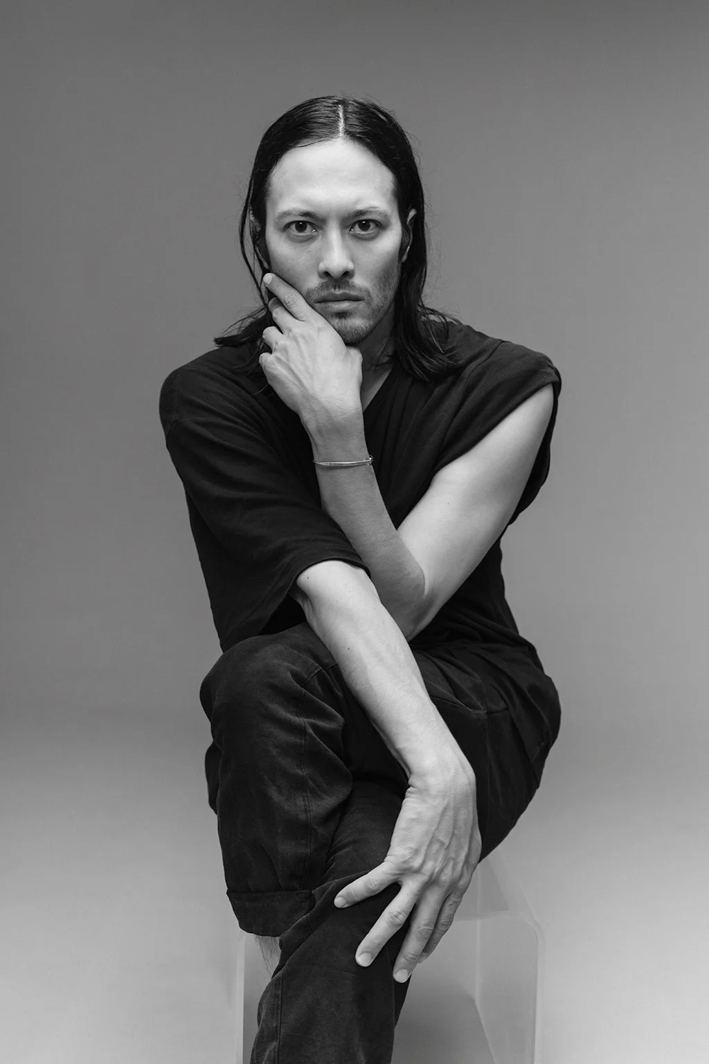 Schwarz-Weiß-Fotografie eines Mannes mit langem dunklem Haar, der gegen eine graue Wand blickt, die Stirn leicht gerunzelt, mit einem Arm auf den Knien und der Hand am Kinn.