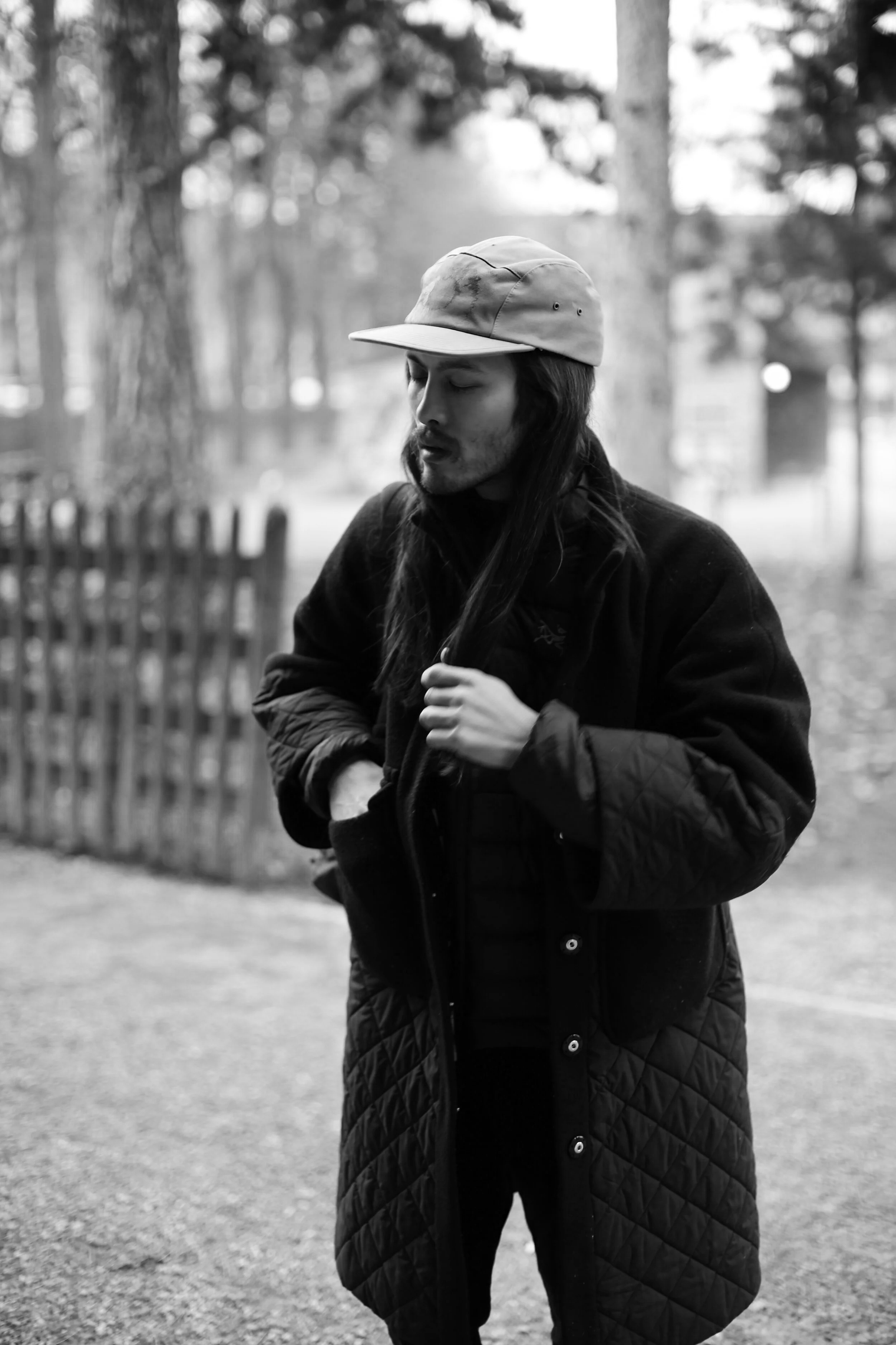 Schwarz-weiß-Foto eines Mannes mit langen Haaren, der eine schwarze Jacke trägt und eine graue Mütze auf hat, steht im Freien vor Bäumen und einem Holzzaun.