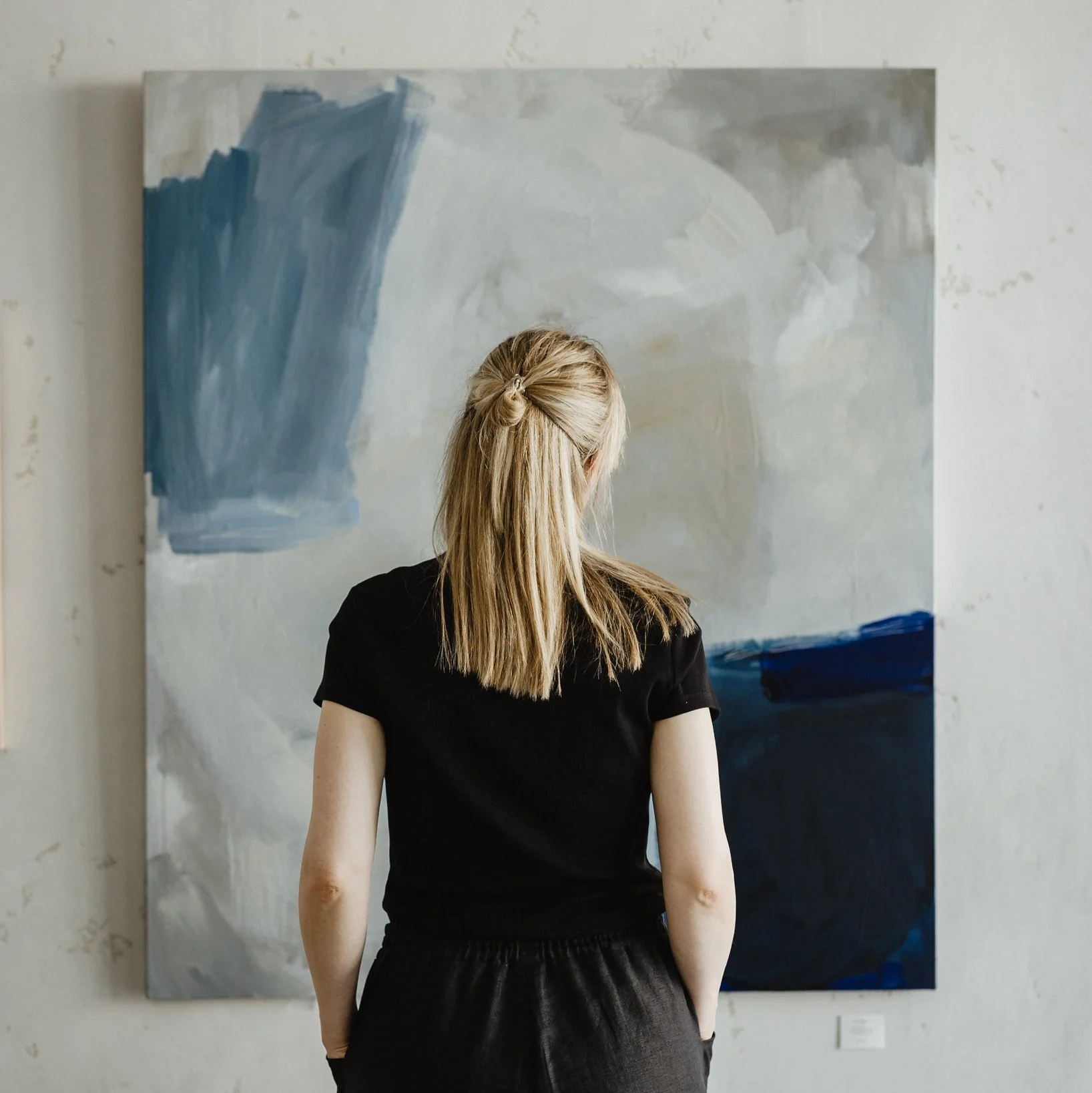 Artist Mareike Böhmer, dressed in black, stands in front of an abstract painting with blue, white, gray, and beige brushstrokes, viewing the artwork in an art gallery.