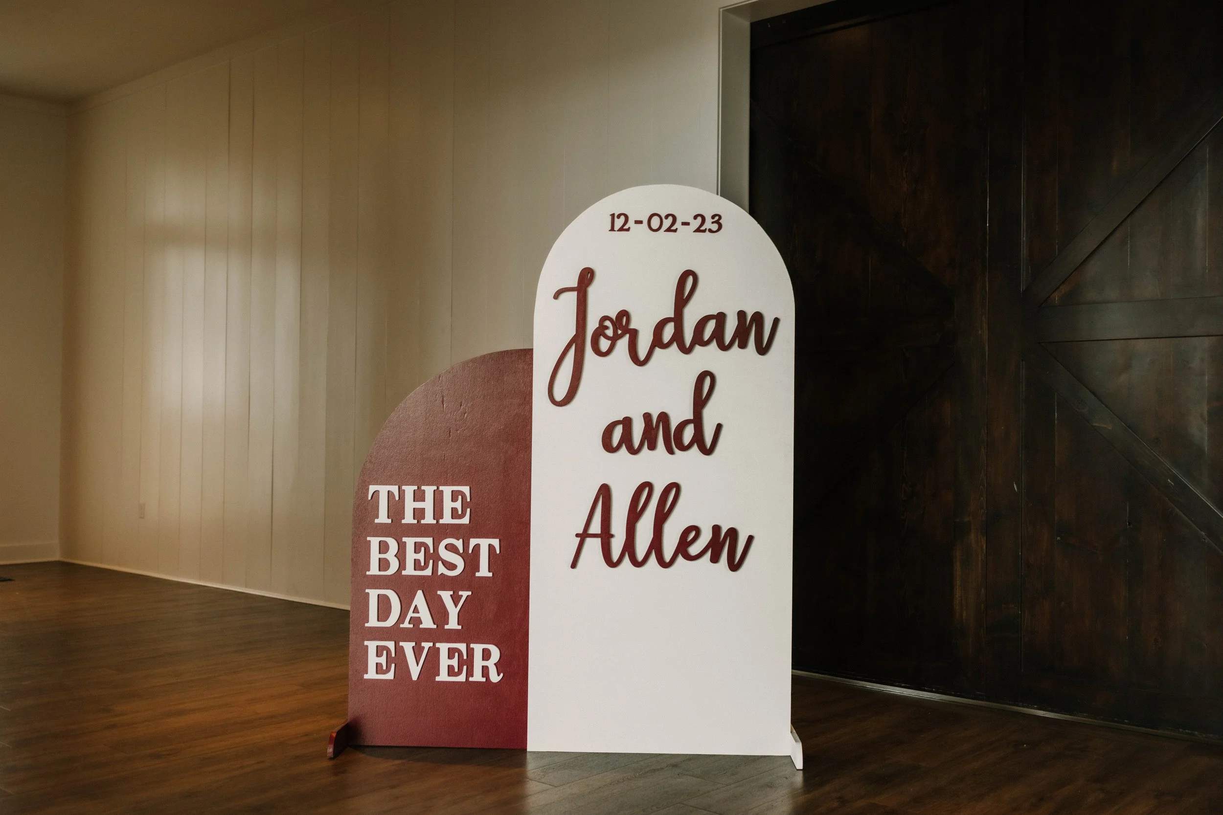 Wedding sign with date 12-02-23 reading "Jordan and Allen" in large cursive and "The Best Day Ever" in bold letters on a wooden floor near dark wooden doors.