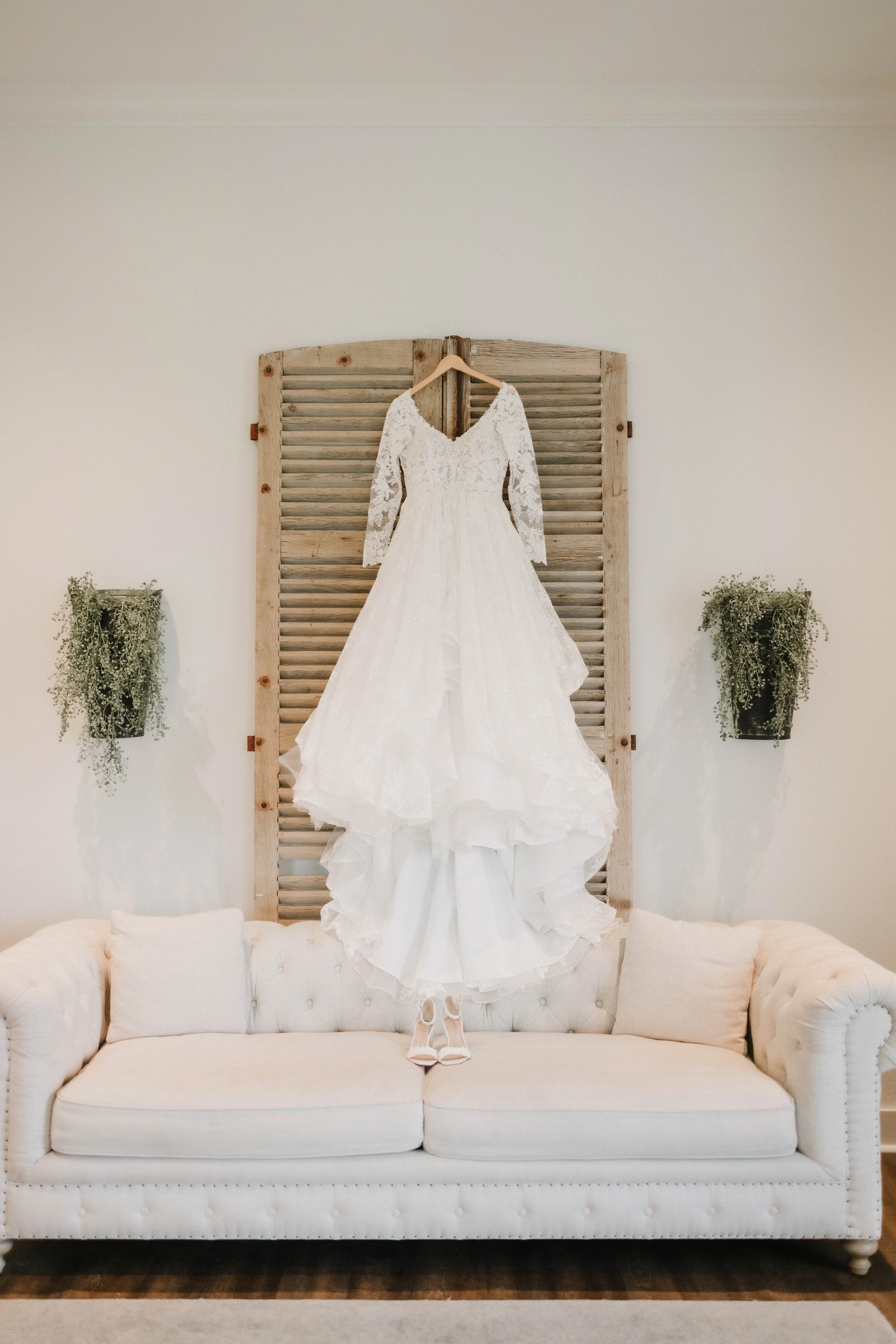 A white wedding dress with lace long sleeves hanging on a wooden headboard, with a pair of white shoes beneath on a light-colored sofa, on a neutral wall with two green potted plants on either side.