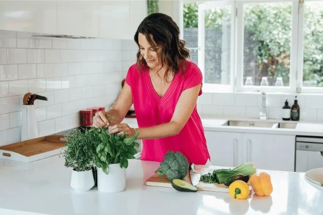 NZs leading dietitian and nutritionist, angela berrill in a pink top, in a white kitchen picking herbs