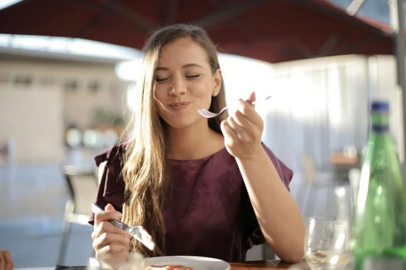 Young woman with long hair enjoying a meal outdoors, holding a fork and smiling with eyes closed.
