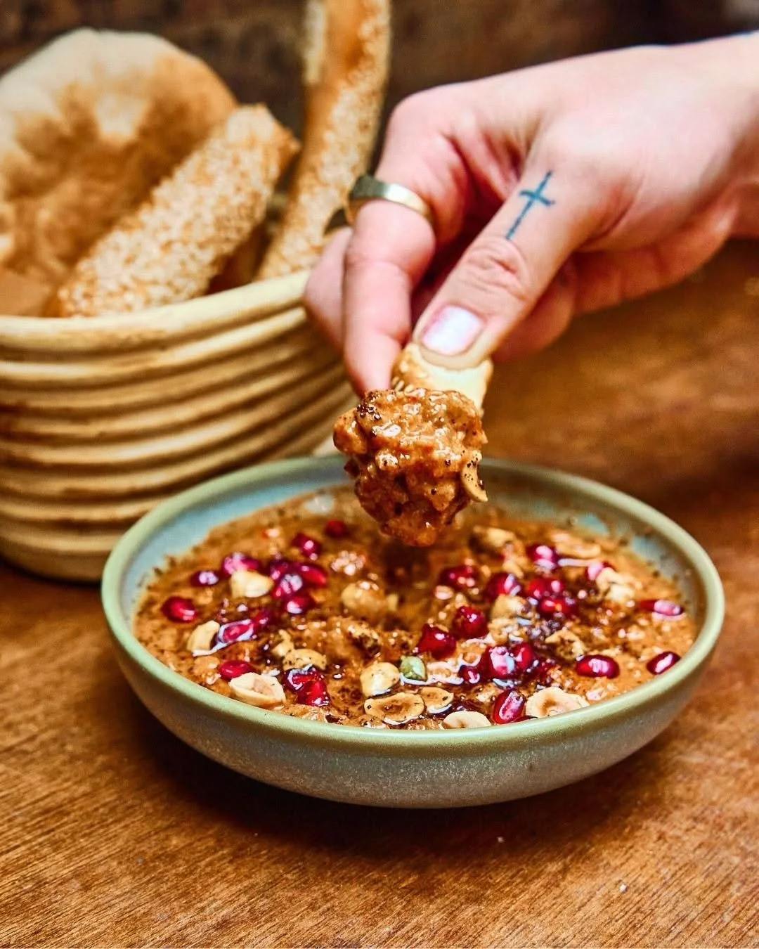 A hand dips bread into a bowl of richly garnished dip topped with pomegranate seeds and nuts,  basket of bread slices in the background on a wooden table—a delicious moment you might encounter on a food tour in Sydney, Australia.
