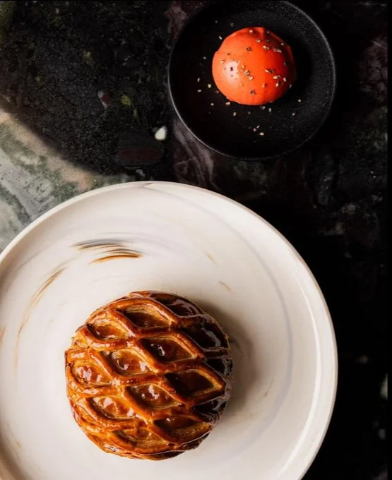 A beautifully presented dish on a marble surface.  The main plate holds a round, golden-brown lattice pastry with a glossy finish. A smaller black dish features a bright orange-red dome-like item sprinkled with seeds.