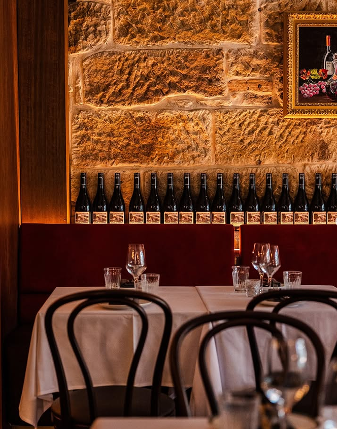 Elegant restaurant interior white tablecloths, wine glasses, dark wooden chairs, and a row of bottles against sandstone wall adorned with framed artwork
