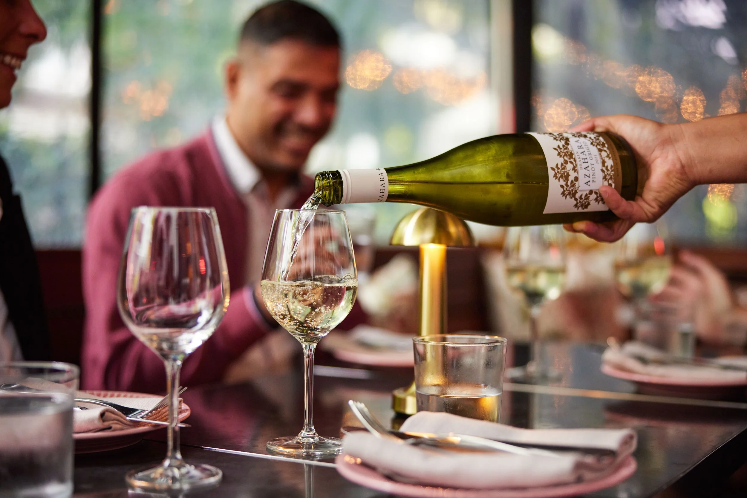 A hand is pouring white wine from bottle into glass at restaurant table during Potts Point food tour. Two people are seated at the table, one of them smiling. The table has another wine glass, rolled napkins, a gold lamp, and a glass of water.