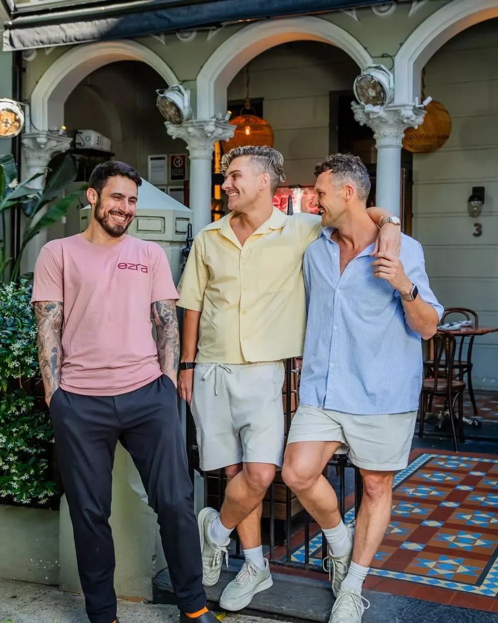 Three men stand smiling outside Ezra restaurant Potts Point, Sydney. Two are leaning against each other, with one man’s arm around the other’s shoulder. dressed casually in shorts and shirts, enjoying a friendly moment together