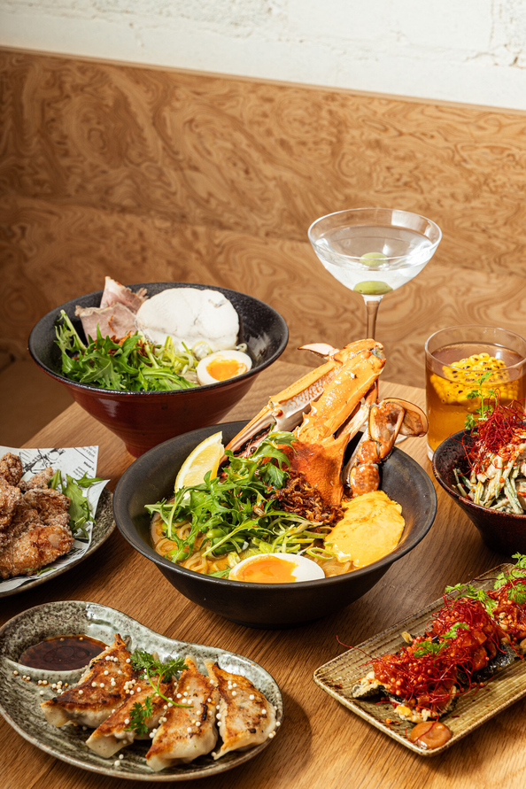 A table set with various Japanese dishes, including ramen with seafood, gyoza, karaage chicken, rice, eggs, and two cocktails arranged on wooden surfaces—captures the vibrant flavors on a food tour in Sydney’s Potts Point.
